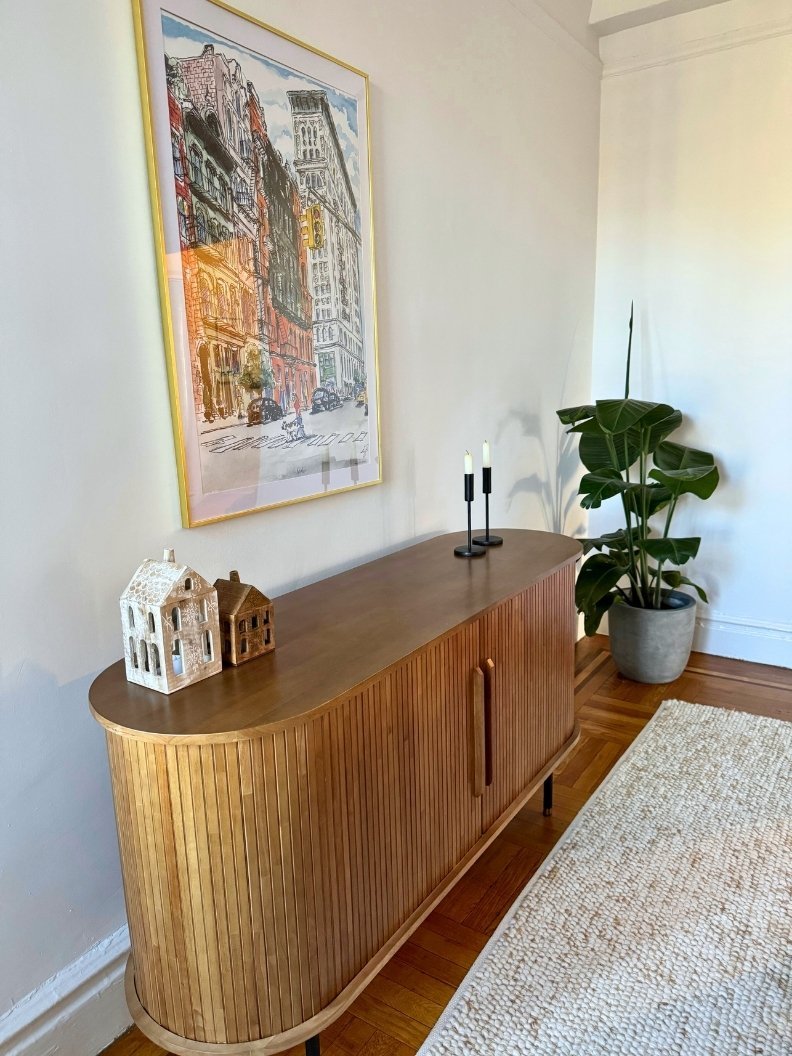 Close-up of a vintage-inspired sideboard with fluted wood detail and curved sides. A large watercolor painting of NYC streets hangs above, with ceramic houses and an indoor plant completing the vignette.