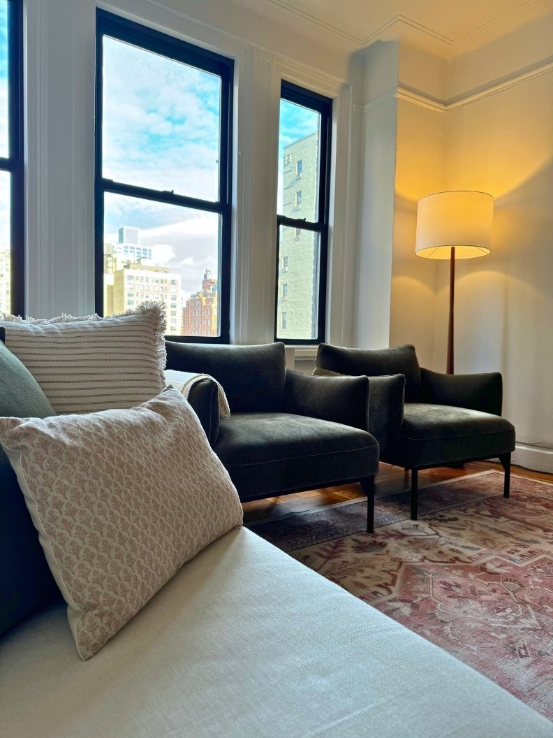 Close-up view of two olive green velvet chairs with textured pillows, a mid-century modern floor lamp, and a coral vintage rug in a NYC living room.