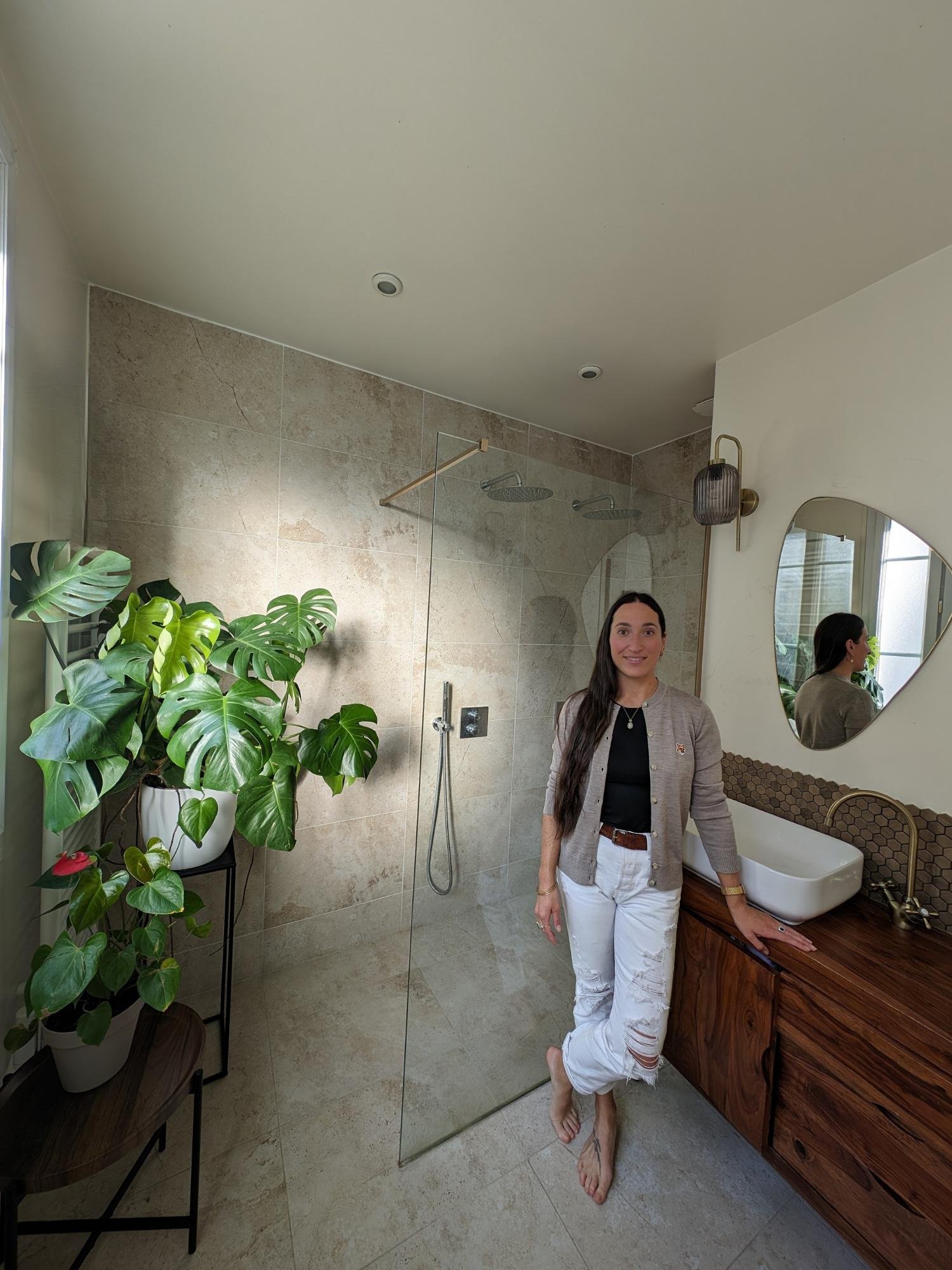 L'Atelier By ATB designer, Alice, standing in a serene, spa-like bathroom with travertine floors, a large monstera plant, and a vintage vanity.