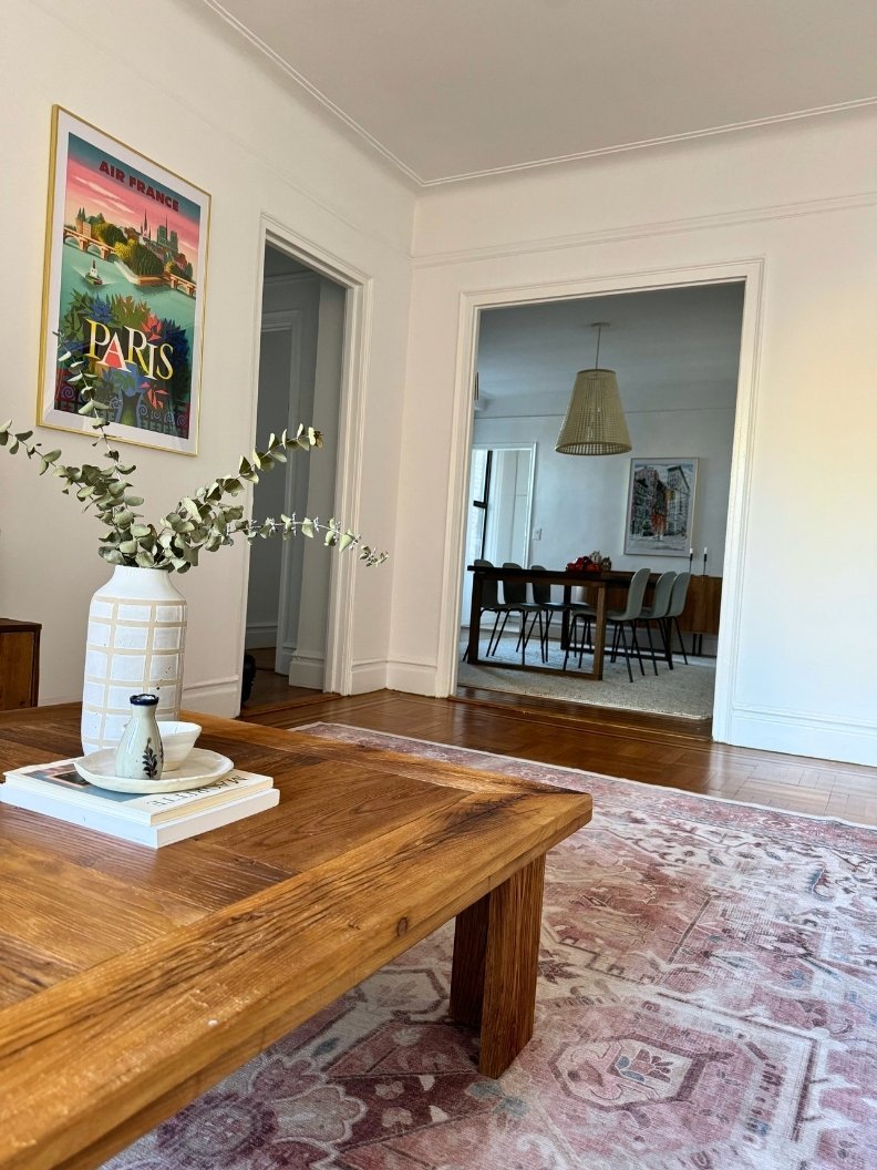 Close-up of a reclaimed wood coffee table with a vintage rug and a vintage Paris advertisement. The dining room is visible in the background.