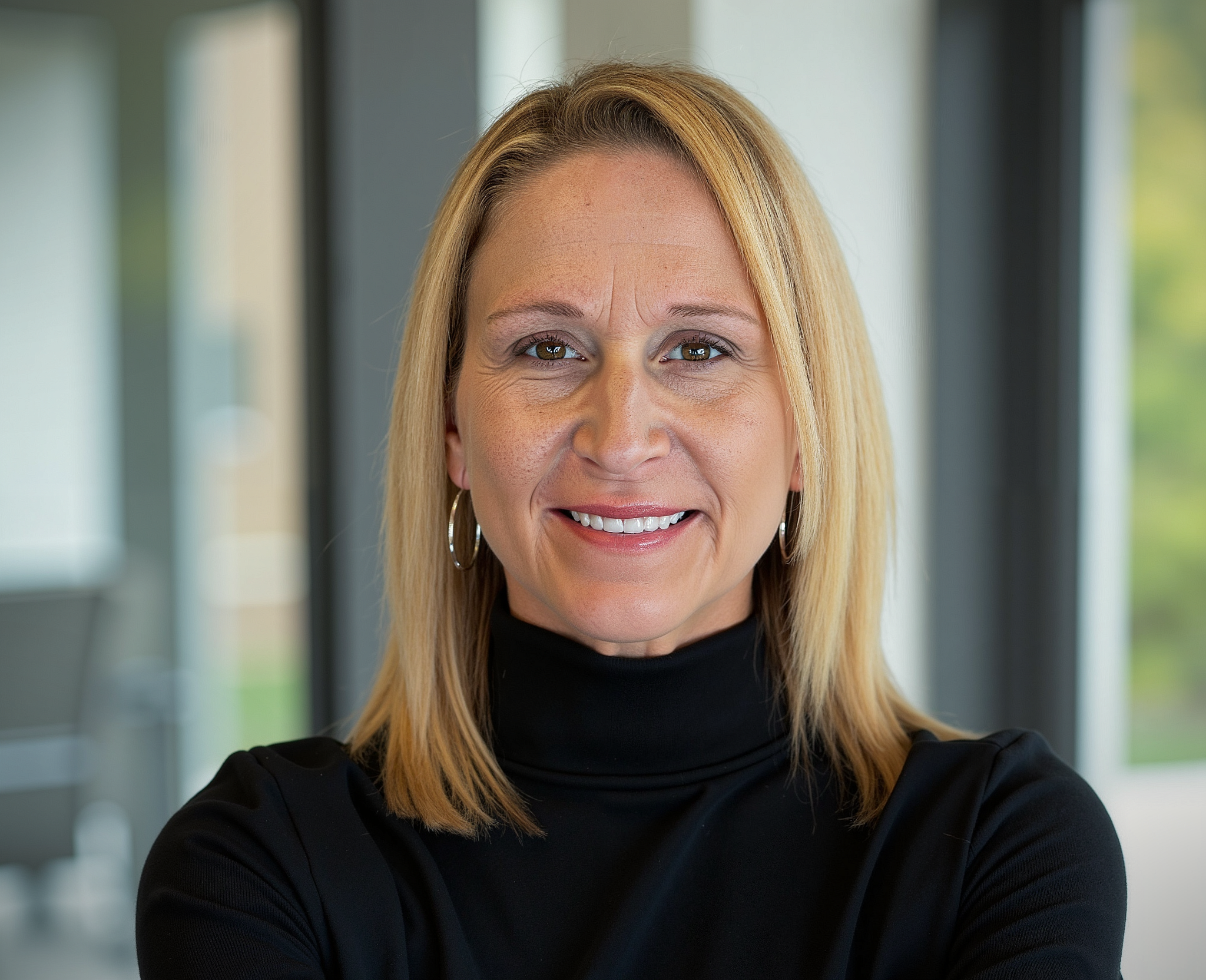 Portrait of a woman with shoulder-length blonde hair, wearing a blue top and a silver necklace, standing in front of a wooden wall.
