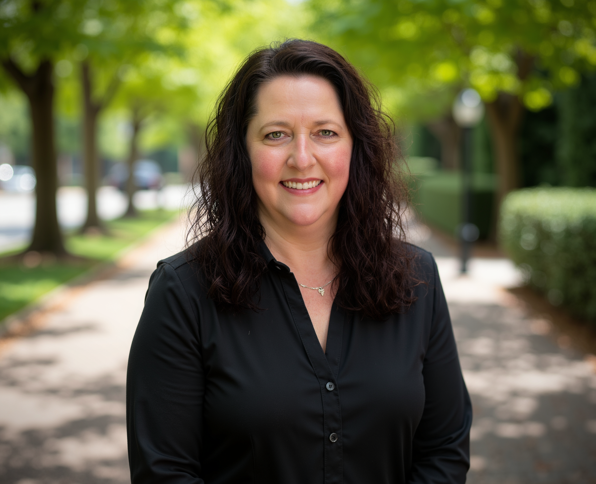 Portrait of a woman with long black hair, wearing a black top, standing in front of a wooden wall.