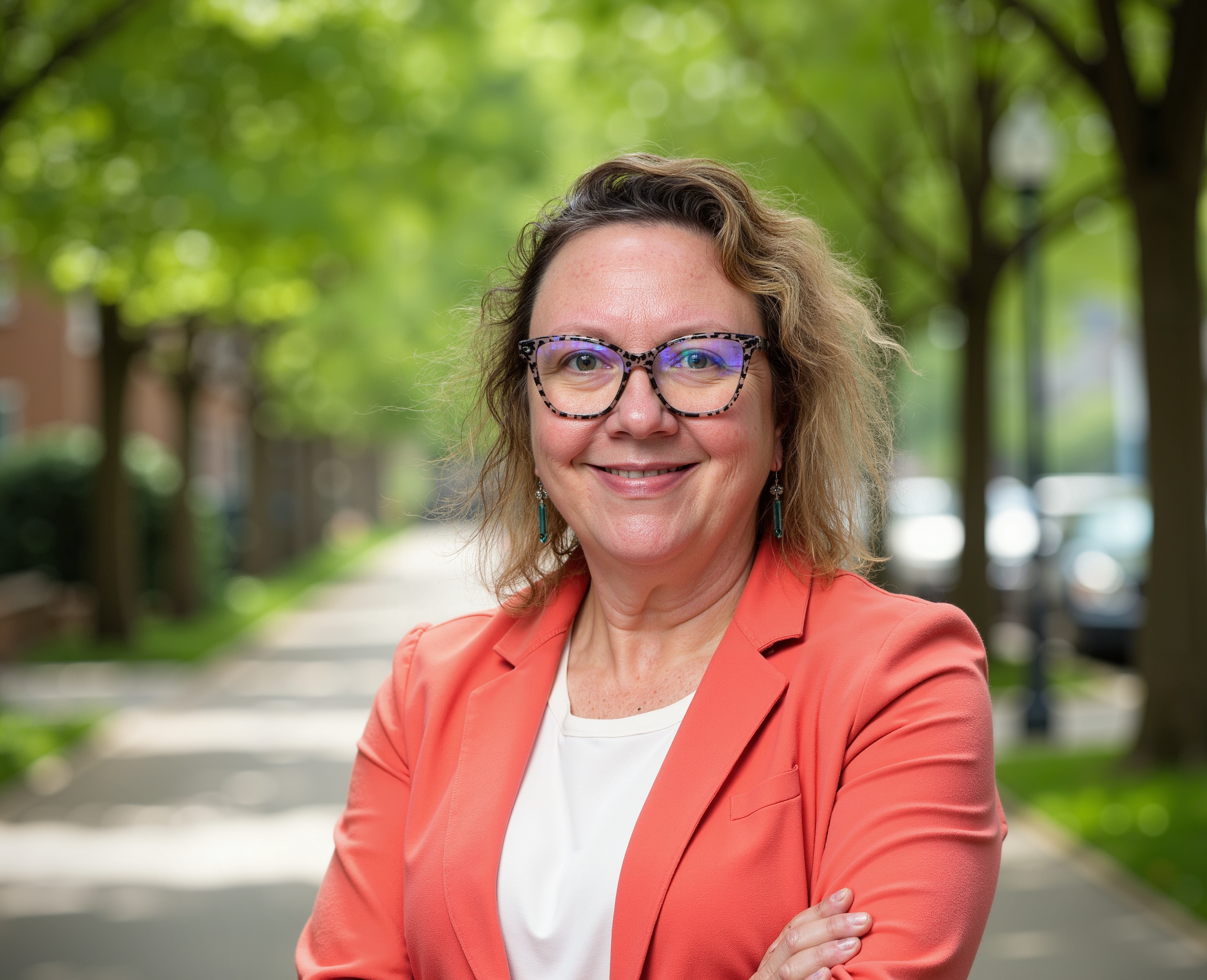 A woman with shoulder-length light brown hair, wearing a green top and a patterned yellow, blue, and white scarf, smiling in front of a wooden background.