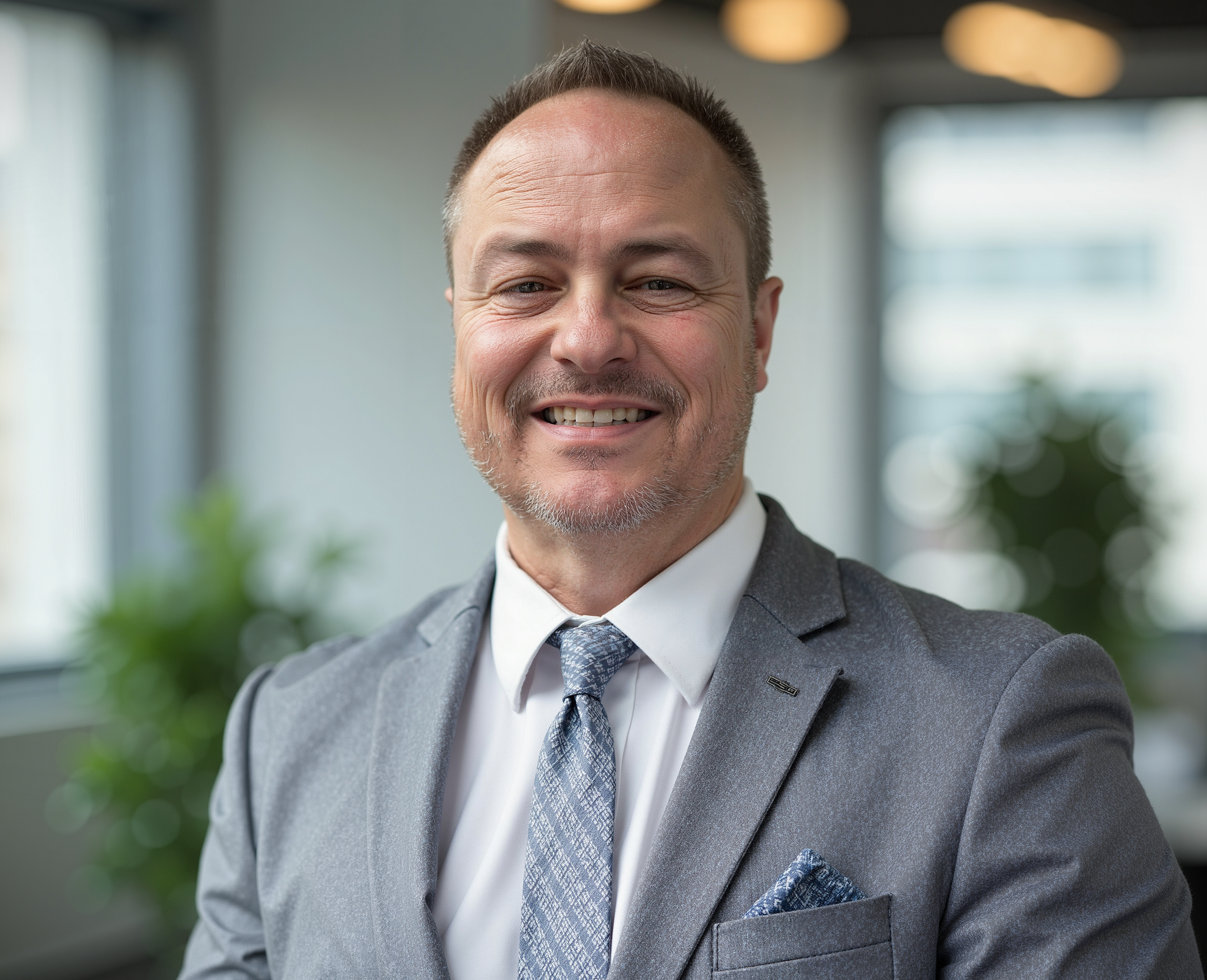 A smiling middle-aged man with blue eyes and short brown hair, wearing a blue polo shirt with a yellow embroidered logo on the chest, standing against a background of brown wooden planks.