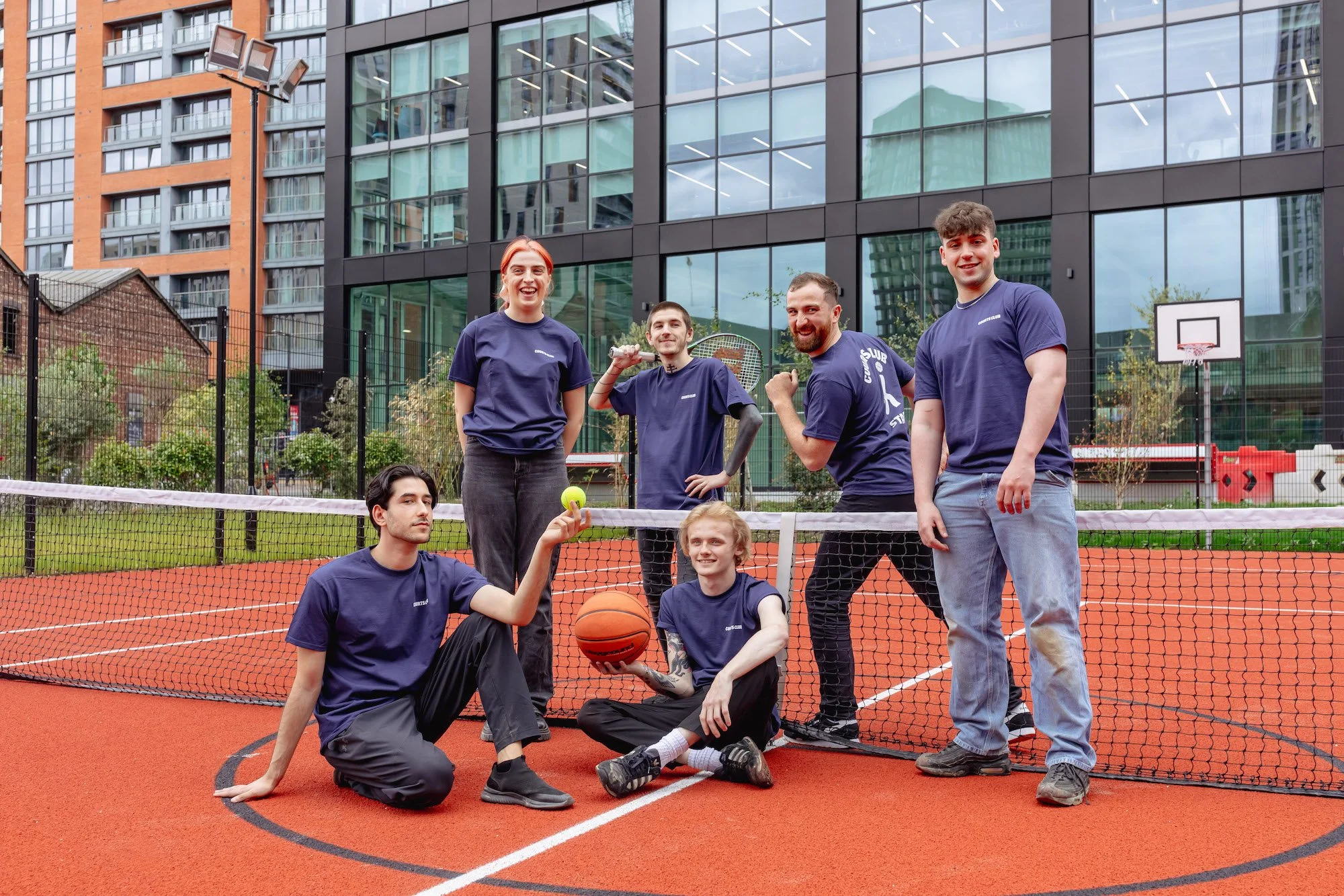 A group of six young adults, five men and one woman, in matching navy blue t-shirts, standing and sitting on a red outdoor tennis or basketball court with a net, surrounded by modern buildings and greenery, holding sports balls and smiling at the camera.