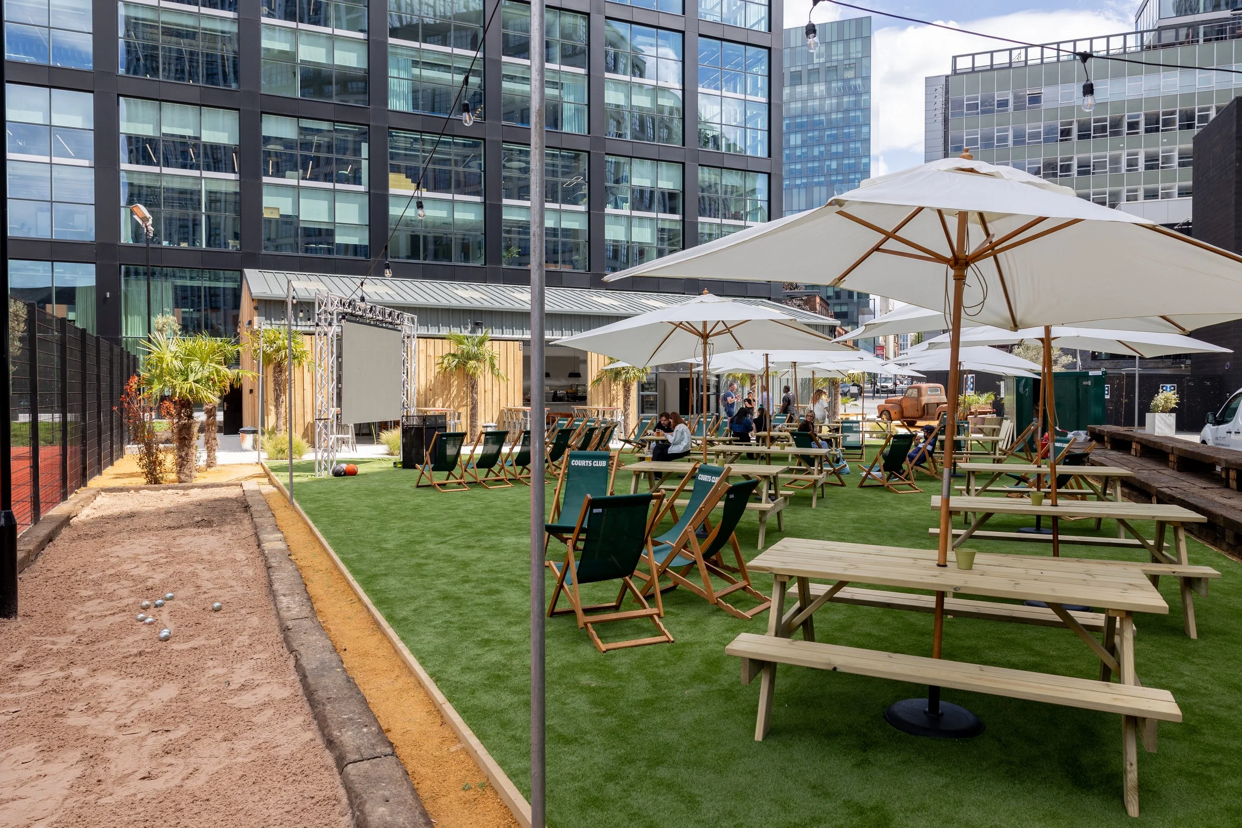 Outdoor urban patio with wooden picnic tables, large white umbrellas, and a small bocce ball court, with people sitting and socializing amidst modern glass buildings.