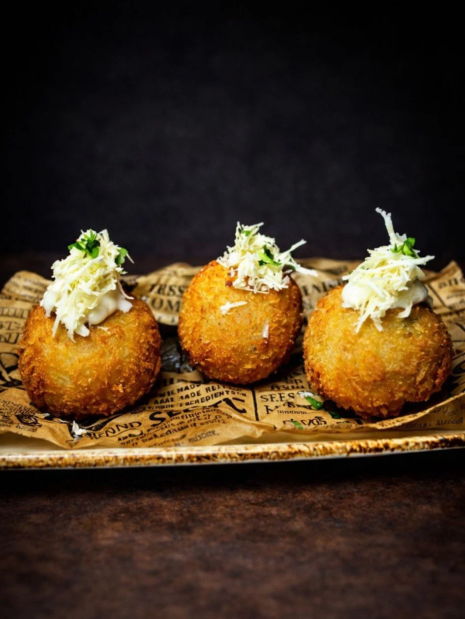 Three breaded, fried ball appetizers topped with shredded cheese and herbs on a paper-lined tray against a black background.