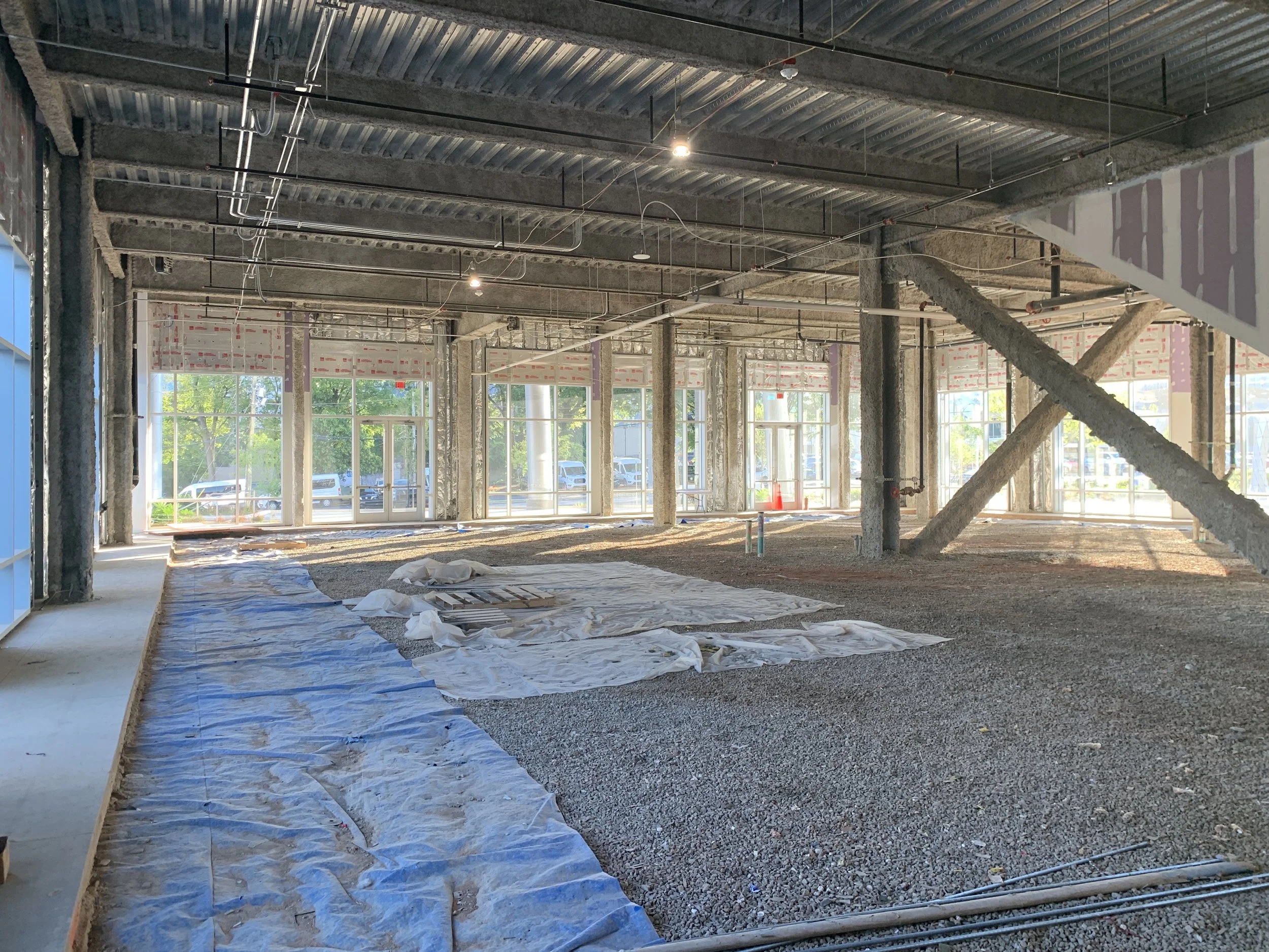 Interior of a building under construction with large windows, exposed ceiling, and gravel floor. Plastic sheets cover parts of the floor; some construction materials are visible.