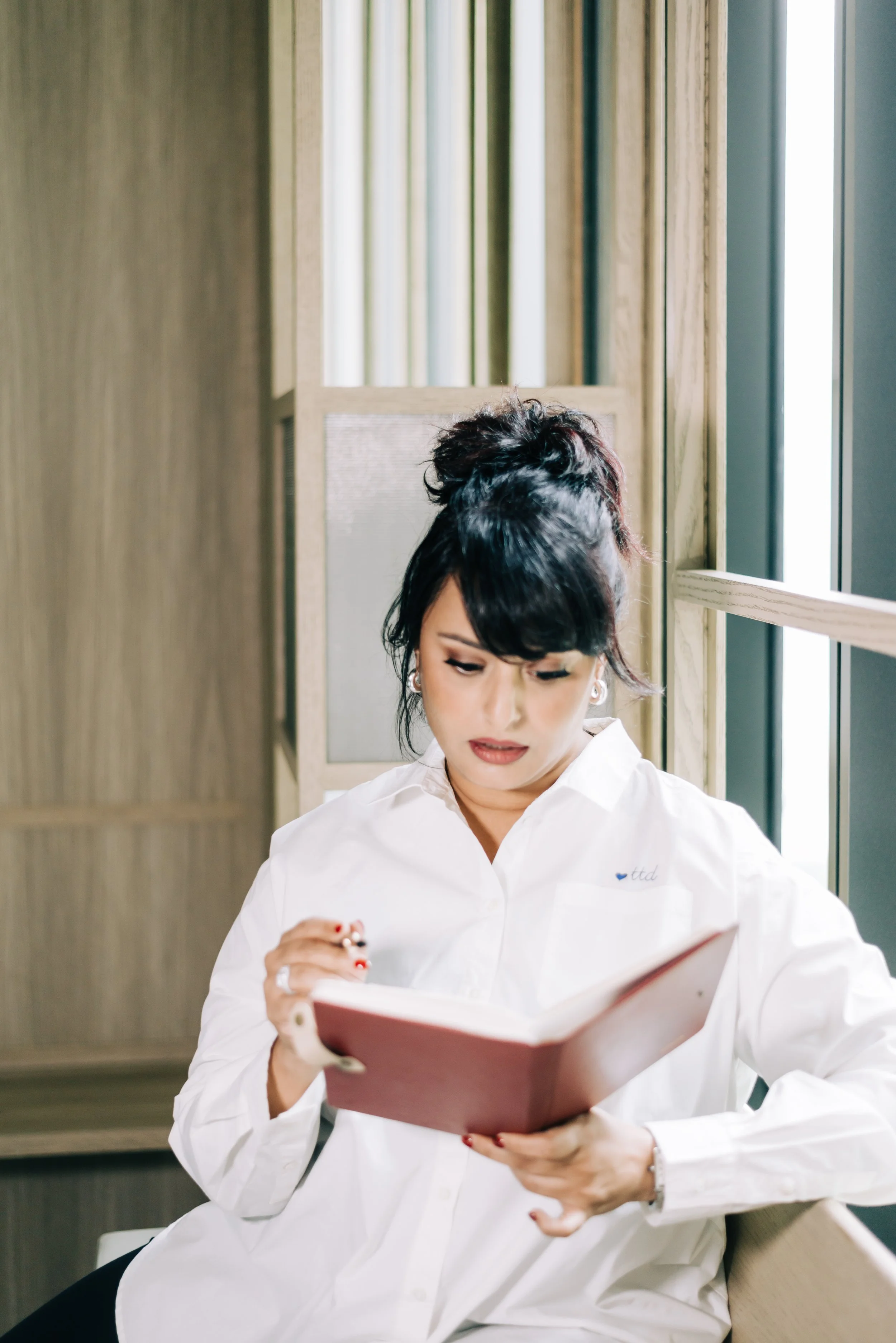 A woman with dark hair styled in an updo, wearing a white shirt, is seated indoors and reading a maroon notebook or menu.