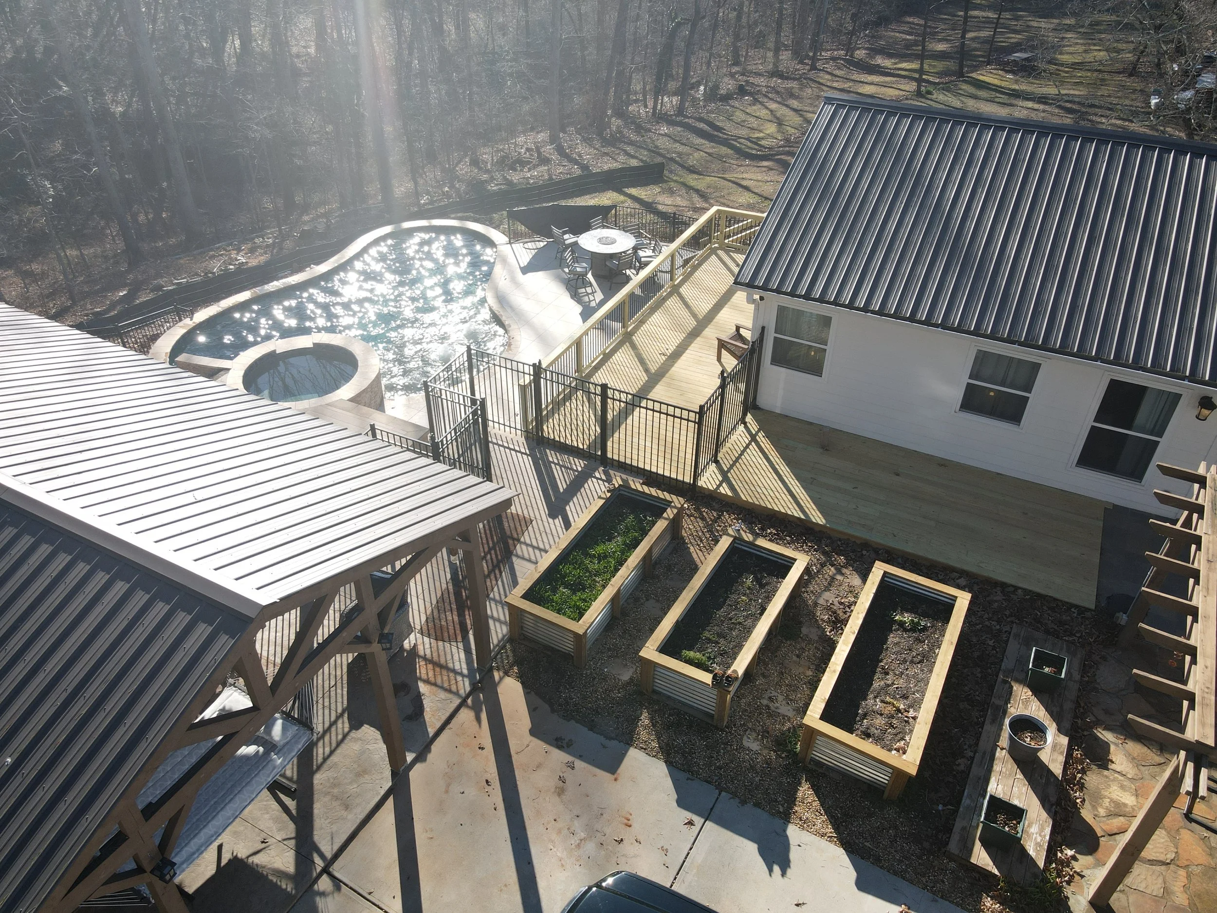 An aerial view of a backyard with a swimming pool, a patio area with a round table and chairs, a house with a metal roof, and wooden garden beds, surrounded by trees.