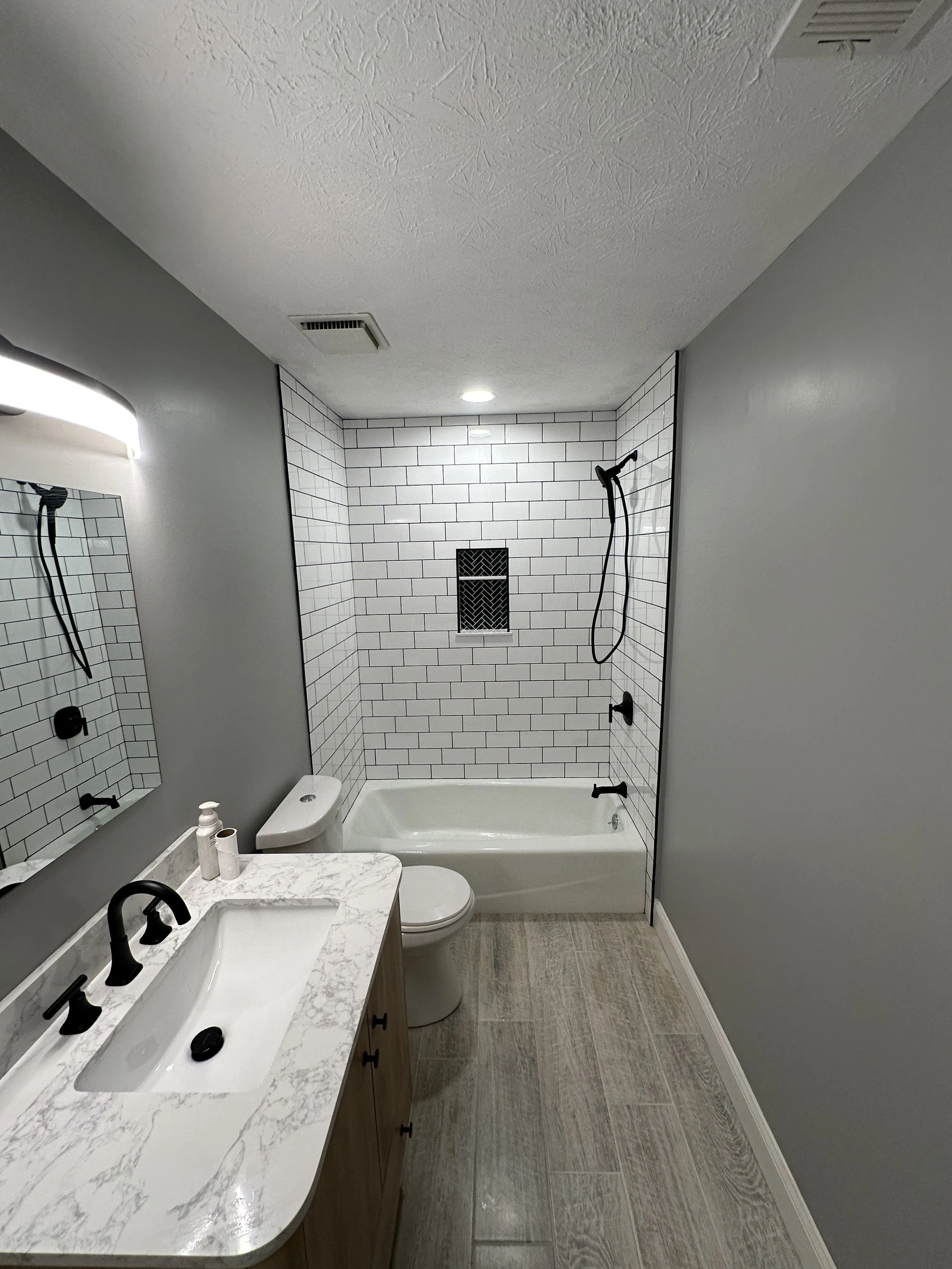 A modern bathroom featuring a white bathtub, a vanity with a marble countertop, a rectangular sink, a mirror, a toilet, and a shower area with white subway tiles and black fixtures.