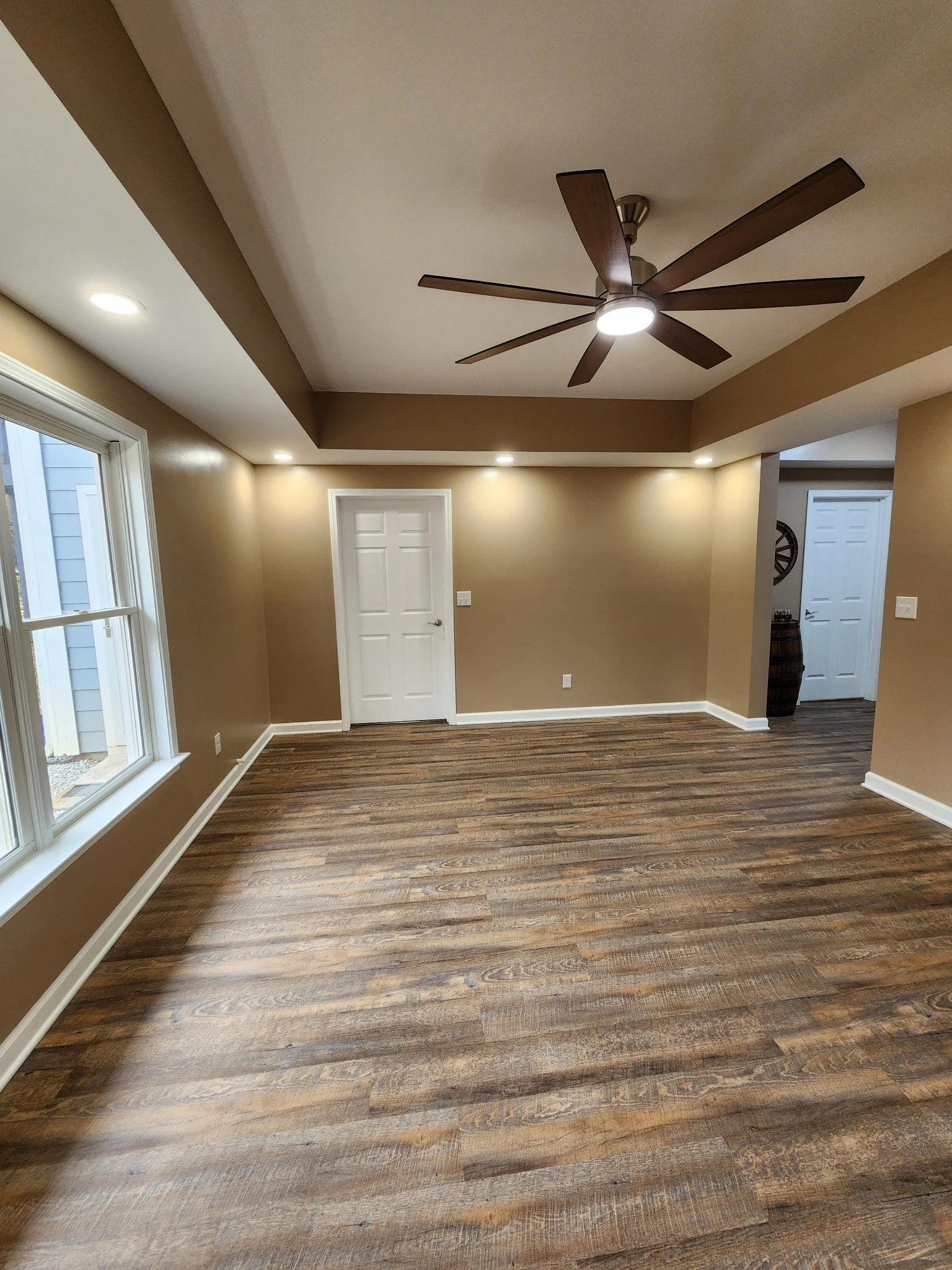 A room with tan walls, natural wood flooring, a white door, a window, white trim, and a ceiling fan with wooden blades and an integrated light.