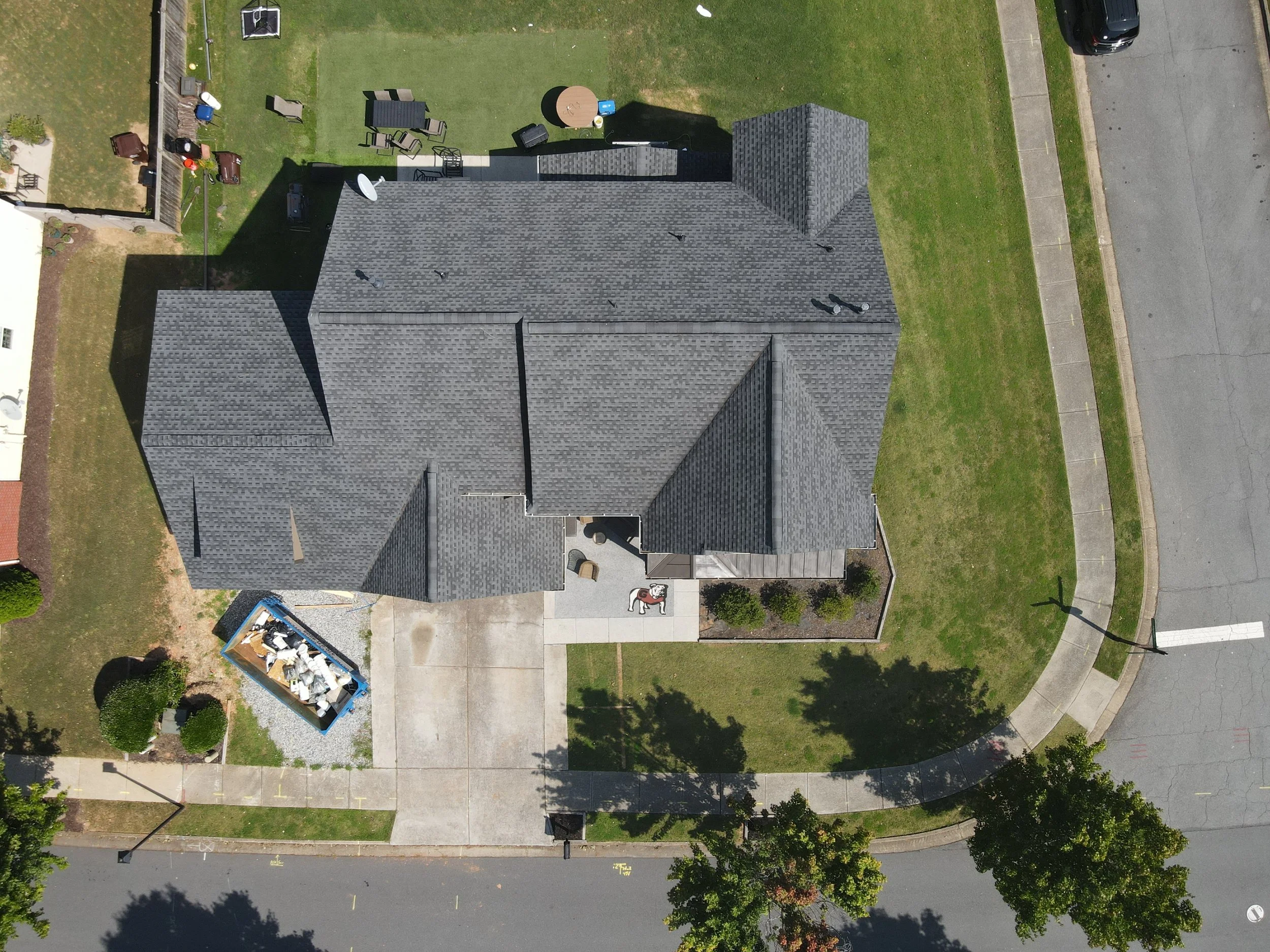 An aerial view of a house with a dark gray roof, a backyard with outdoor furniture, and a lawn surrounded by sidewalks and a street.