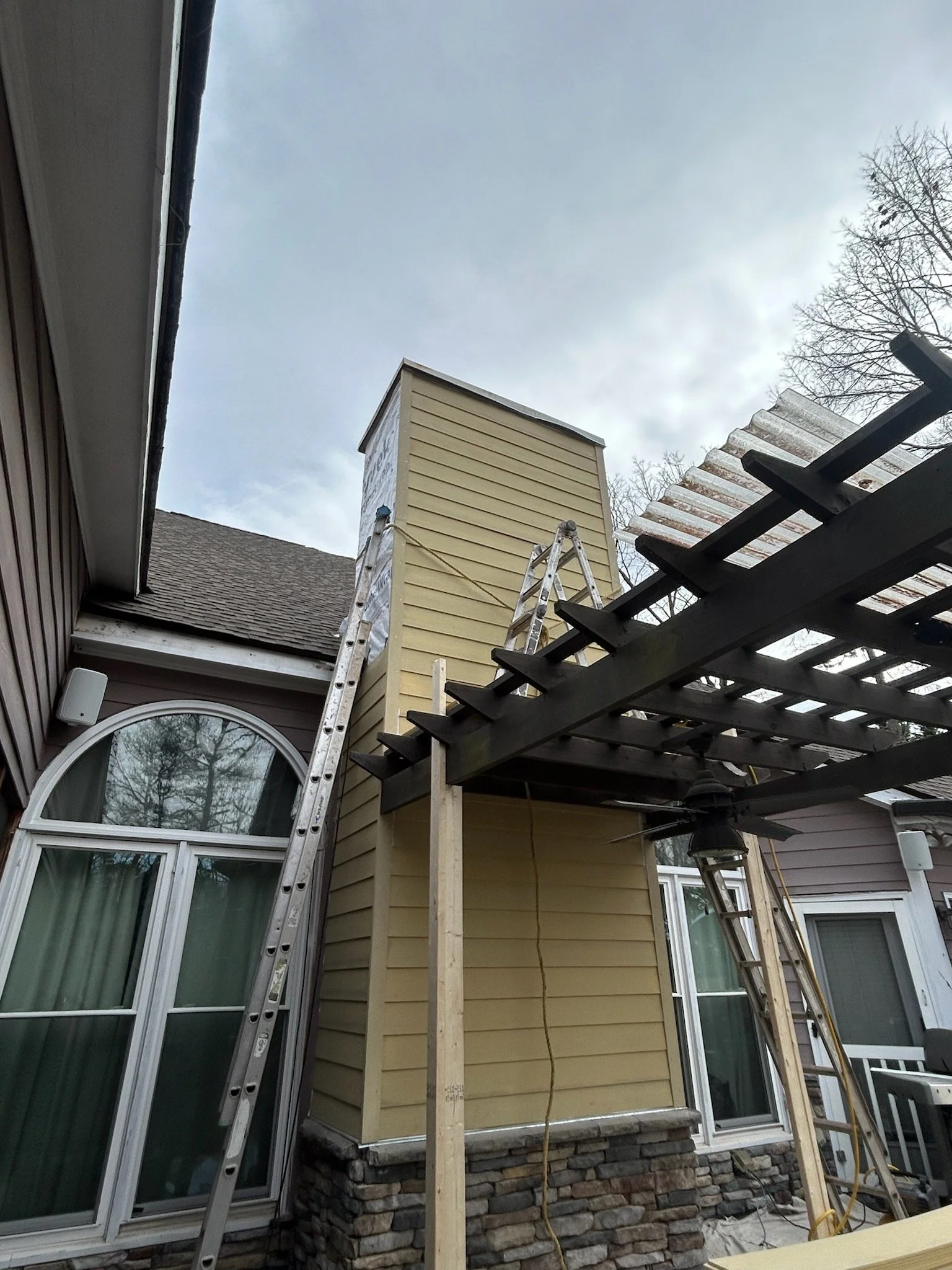 A house exterior with a chimney, a window, and a newly built porch structure with wooden supports and a lattice framework, during construction or renovation.
