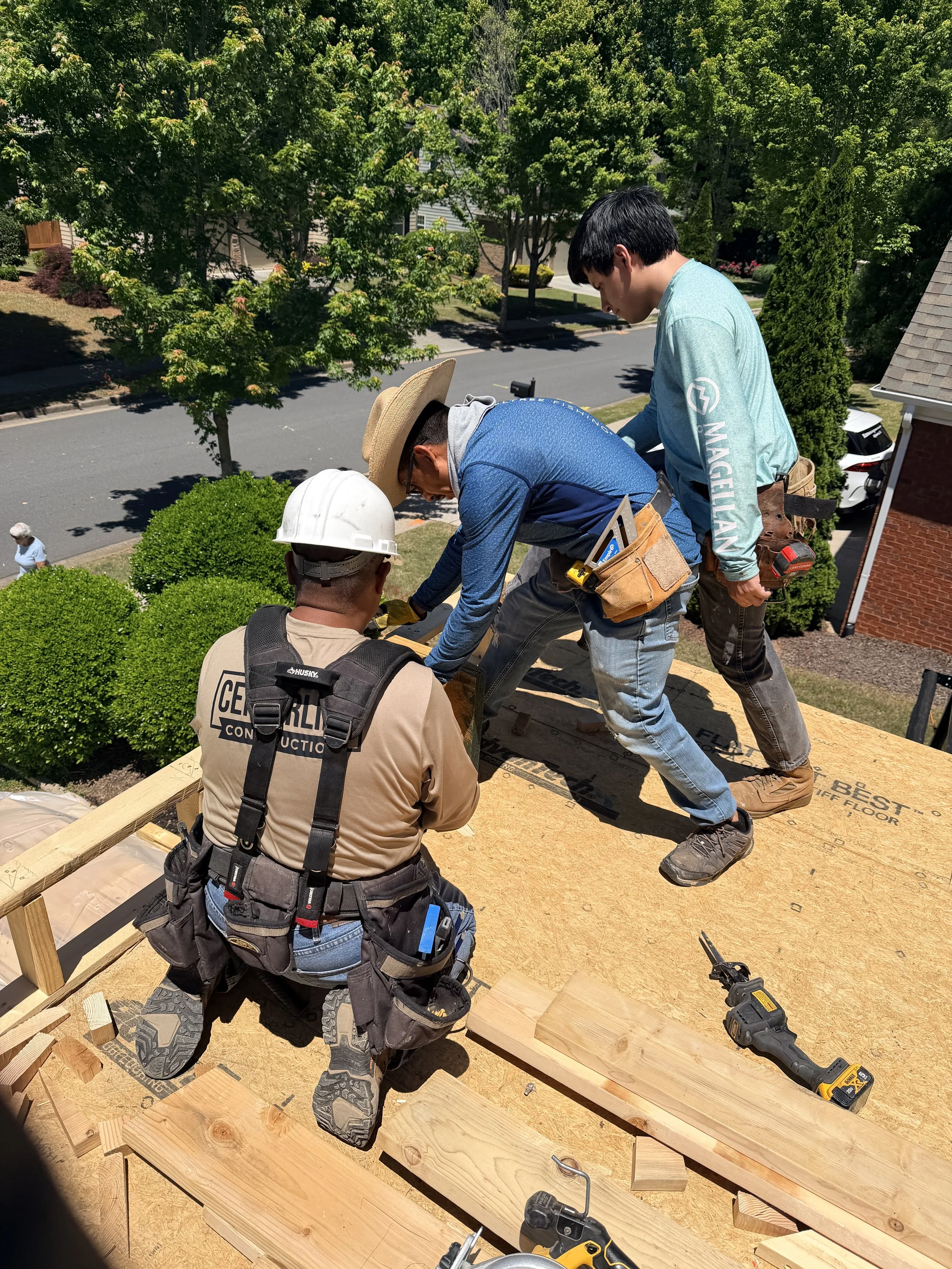 Four workers installing or constructing a wooden structure outdoors on a sunny day, with green trees and a street in the background. They are wearing safety gear and tools.