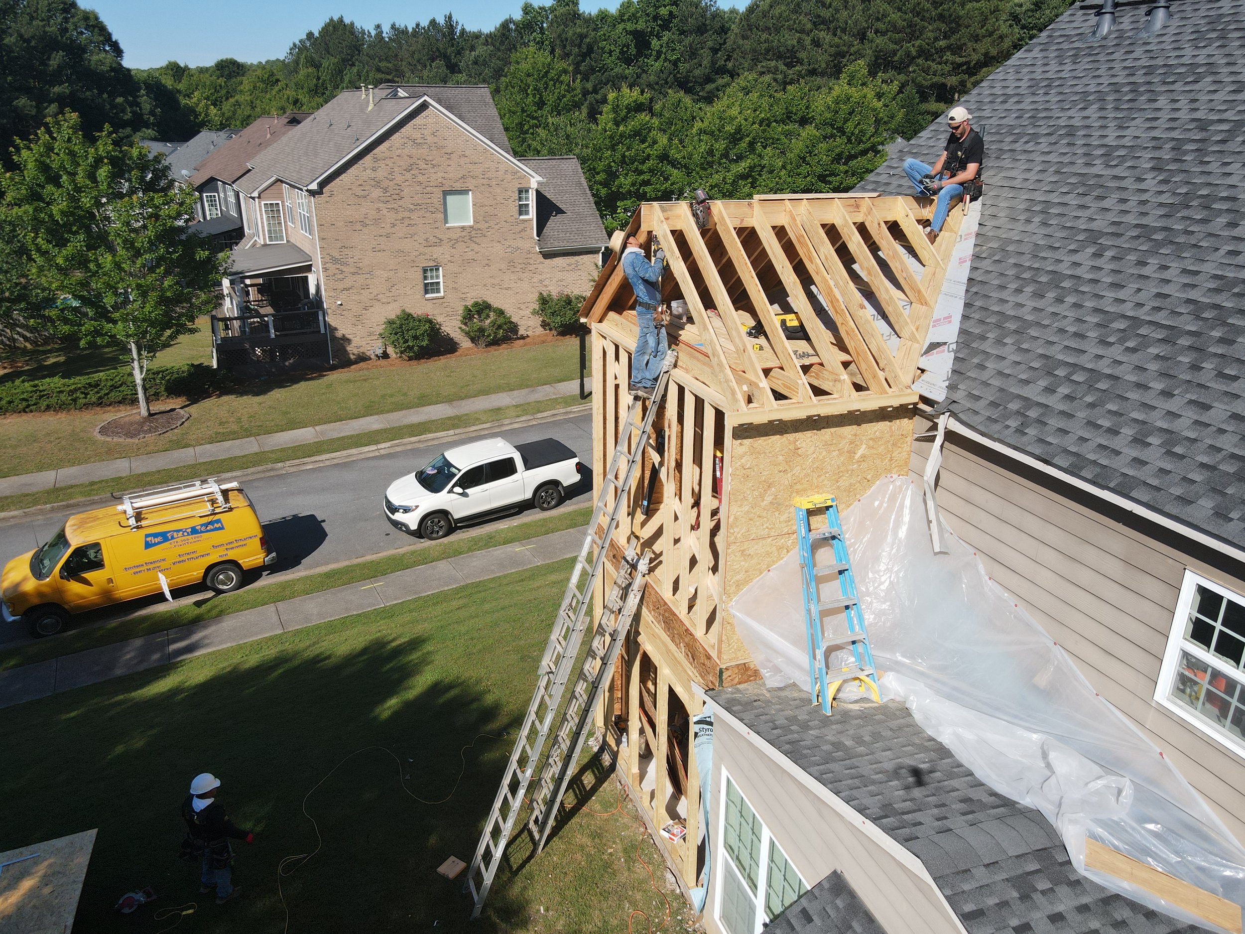 Two construction workers install a wooden roof framework on a house. One worker is seated near the top of the roof, and the other is standing on a ladder. The house is in a residential neighborhood with trees and other houses visible in the backgroun