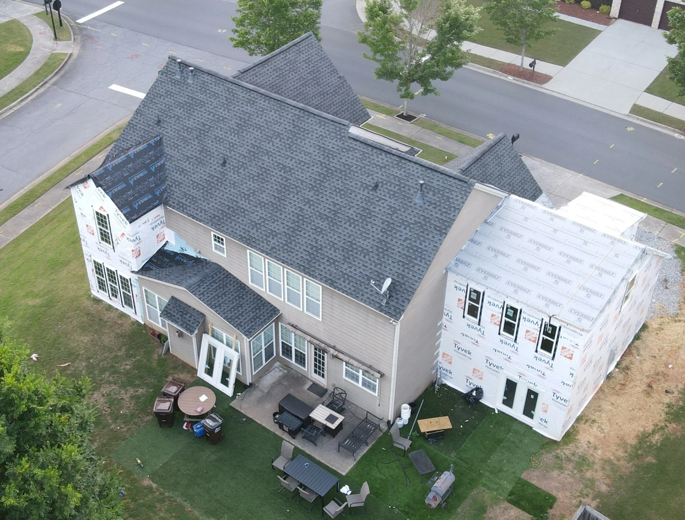 Aerial view of a house under construction with a new roof, surrounded by a lawn, sidewalk, and street, with some outdoor furniture and construction materials visible.