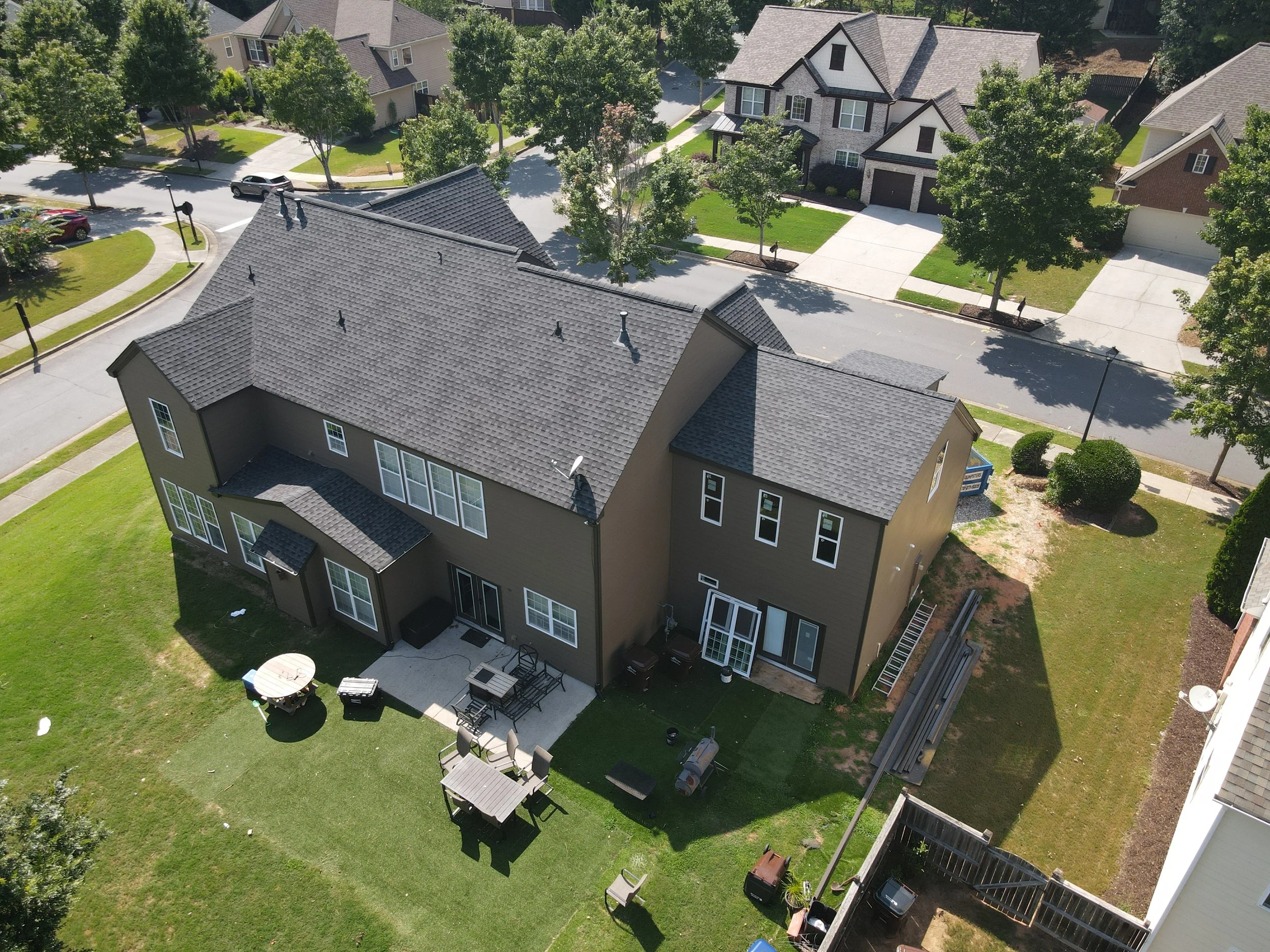 An aerial view of a suburban neighborhood with multiple houses, green lawns, trees, and a curved street with parked cars.