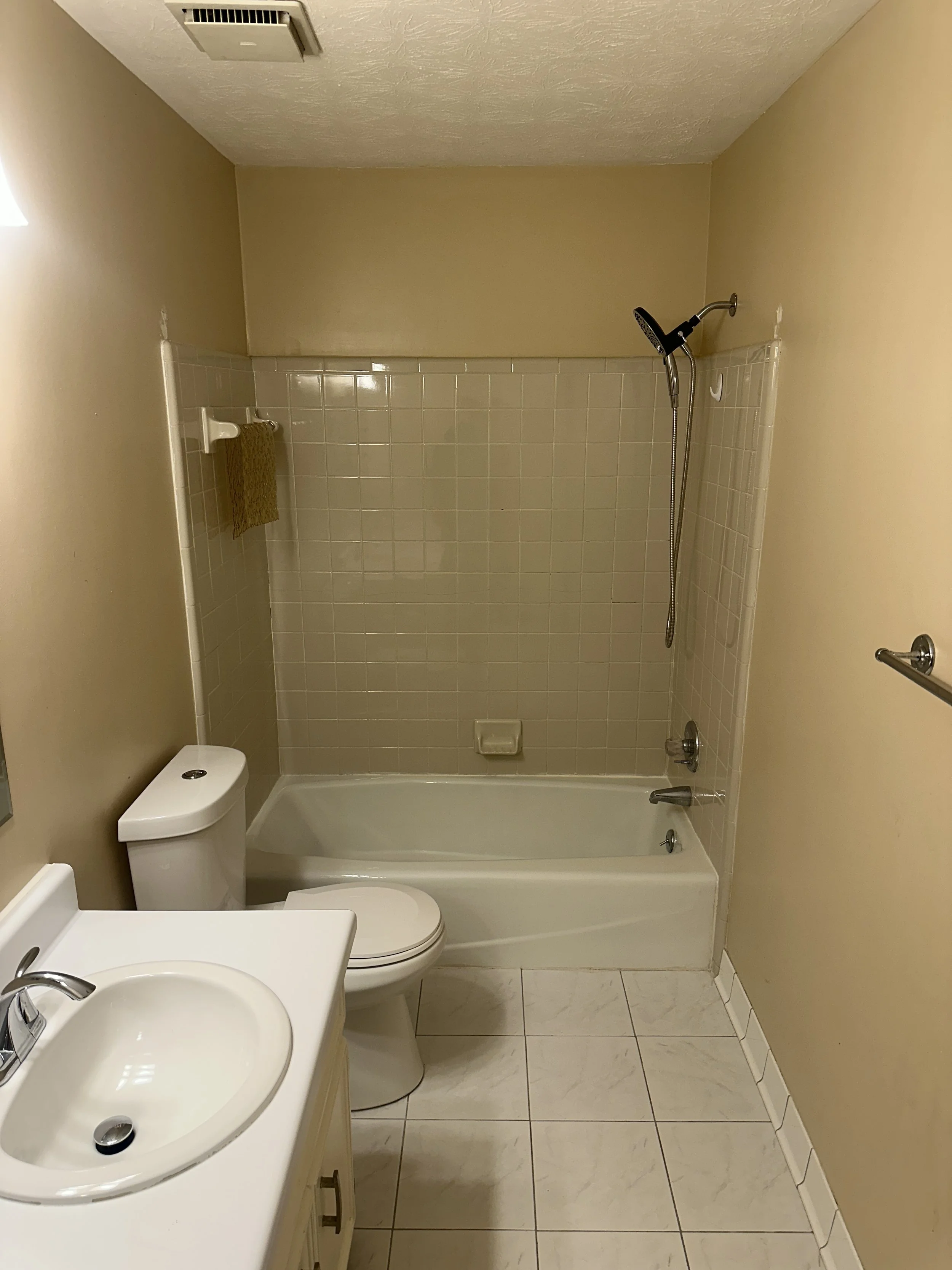 A small bathroom with a beige wall and white tile shower area. There is a yellow towel hanging on the towel bar inside the shower. The bathroom contains a white toilet, a sink with a beige cabinet, and a mirror on the left wall. The floor is tiled wi