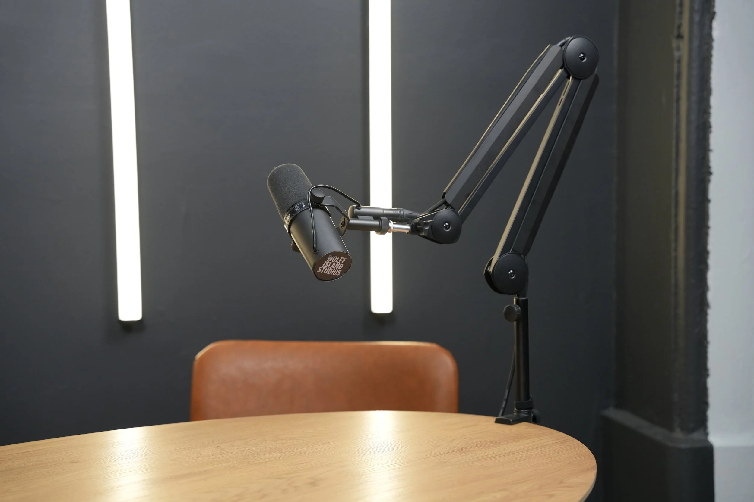 A professional podcasting microphone mounted on an adjustable arm over a wooden table with a brown chair, set against a dark wall with vertical light strips.