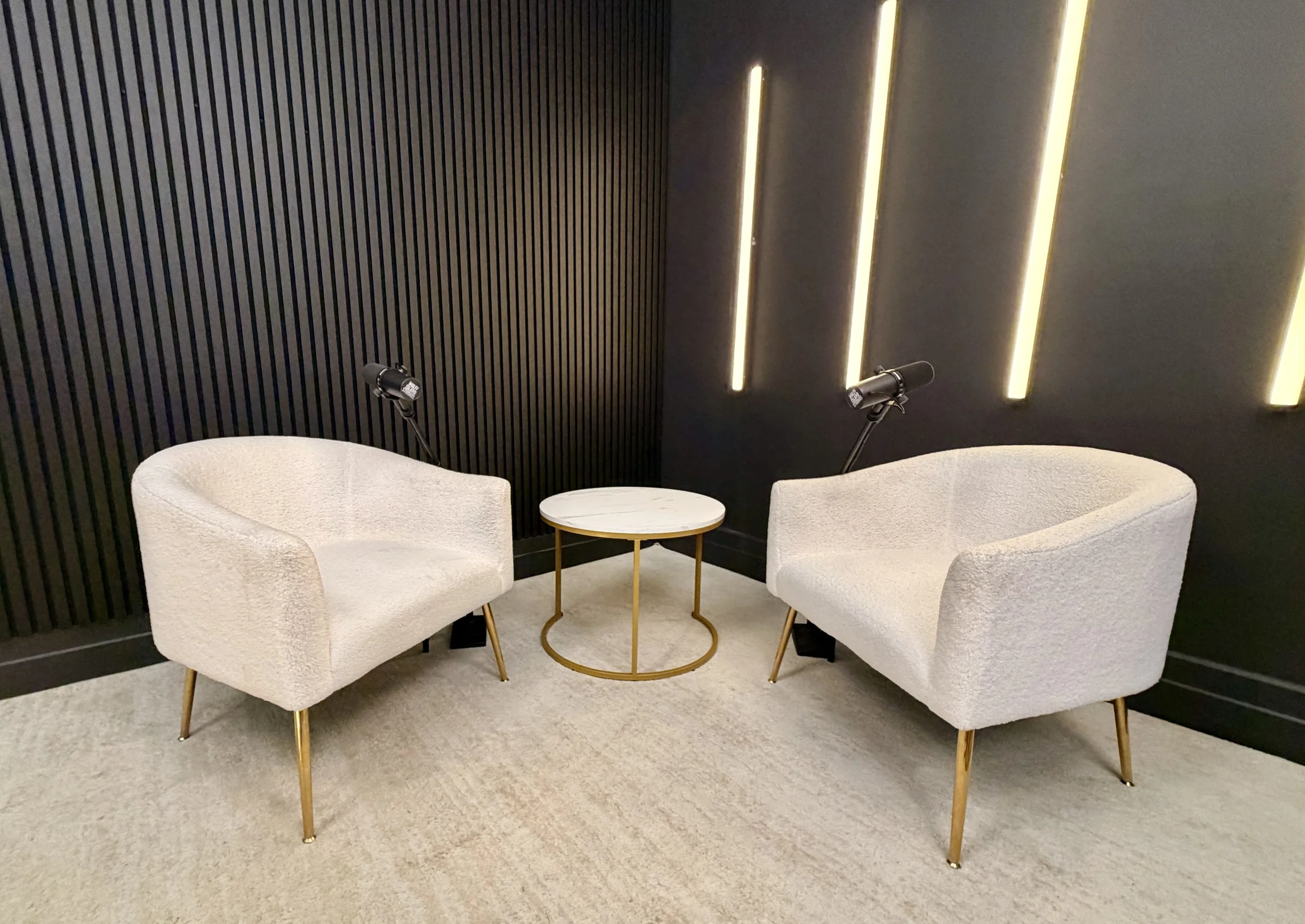 Two white upholstered armchairs with gold legs positioned on either side of a small round table with a marble top and gold frame. Each armchair has a black reading light attached to the wall behind it. The background features a black wall with vertical wooden slats on the left side and three vertical slim light fixtures on the right side, creating a modern, minimalist aesthetic.