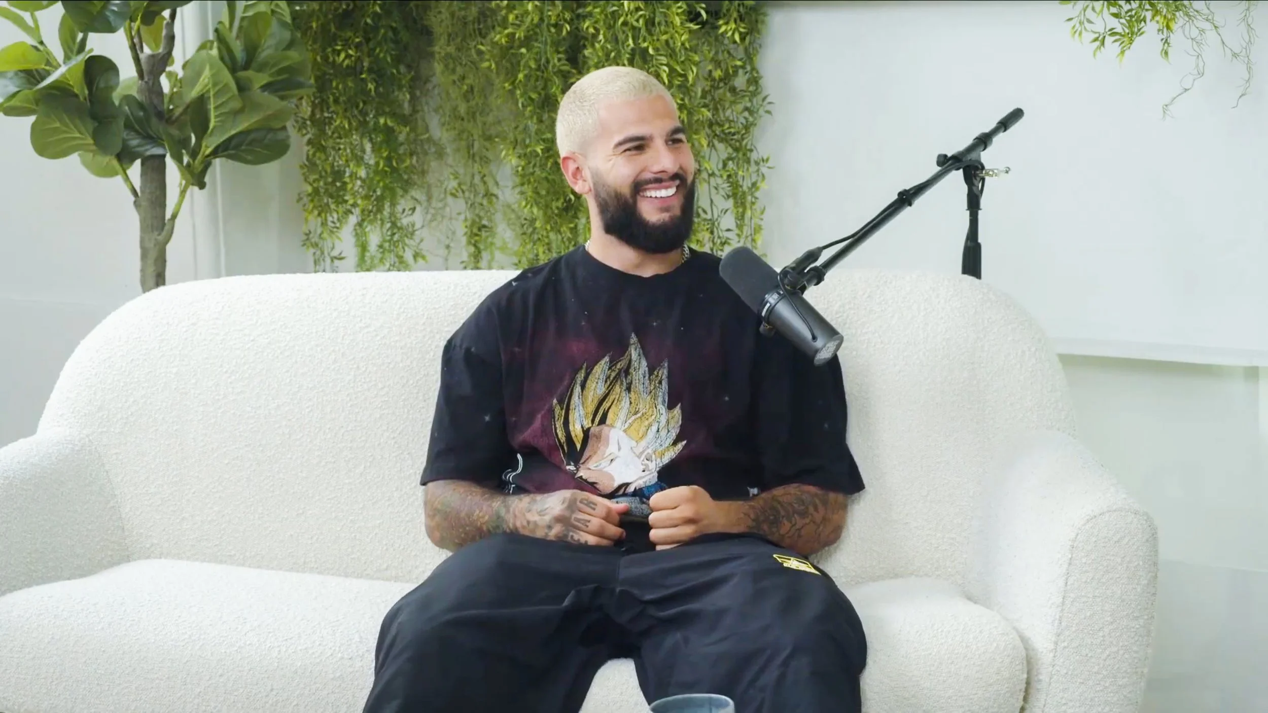 A man with a blonde buzz cut and beard, wearing a black T-shirt with a dragon ball graphic, sitting on a white sofa with a microphone in front of him, smiling and holding a phone. There are green plants and hanging vines in the background.