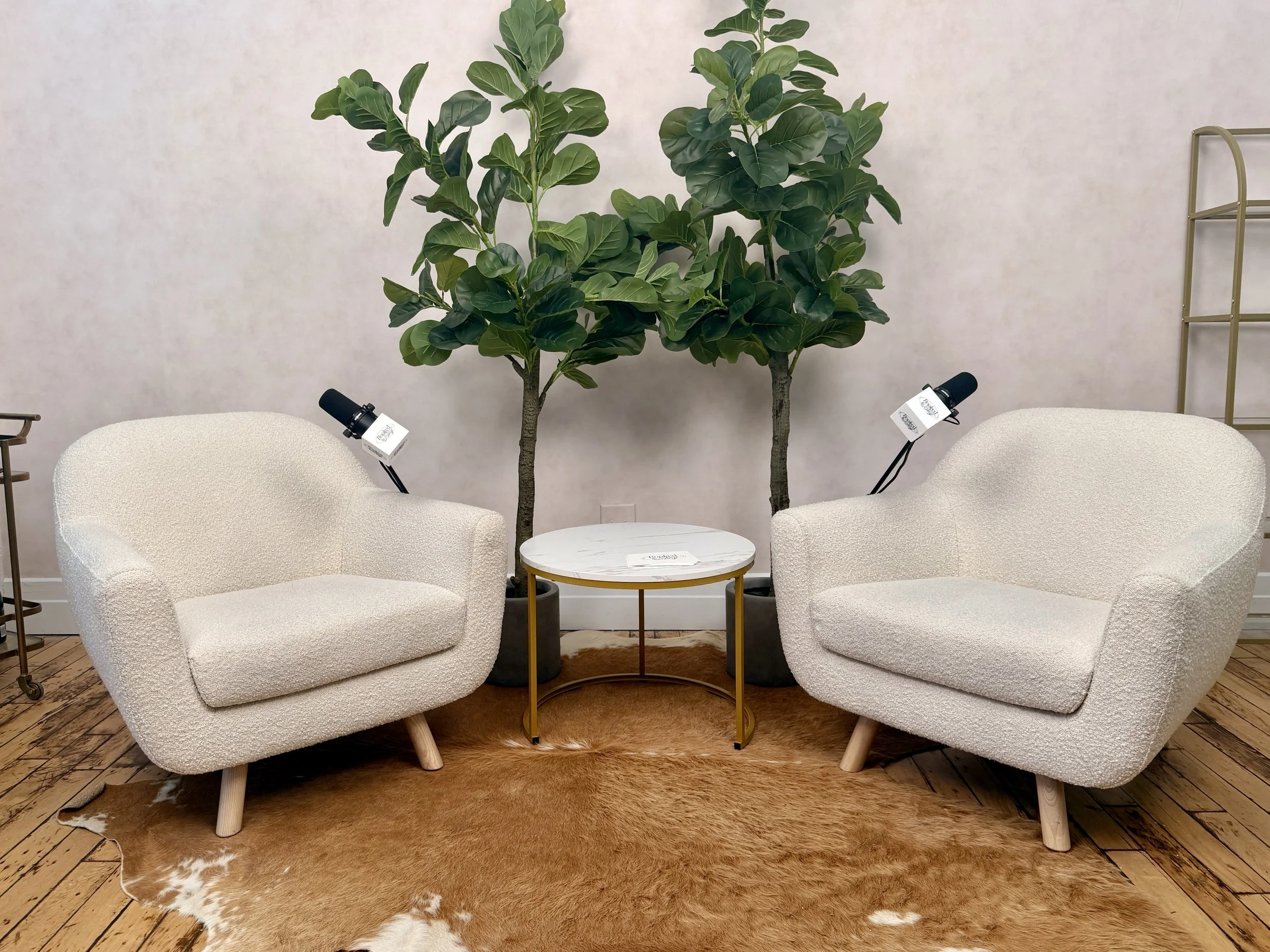 Two cream-colored armchairs with textured fabric and wooden legs face each other, separated by a small round table with a marble top and gold legs, on a cowhide rug. Behind the chairs are two large potted plants with broad green leaves. A beige wall 