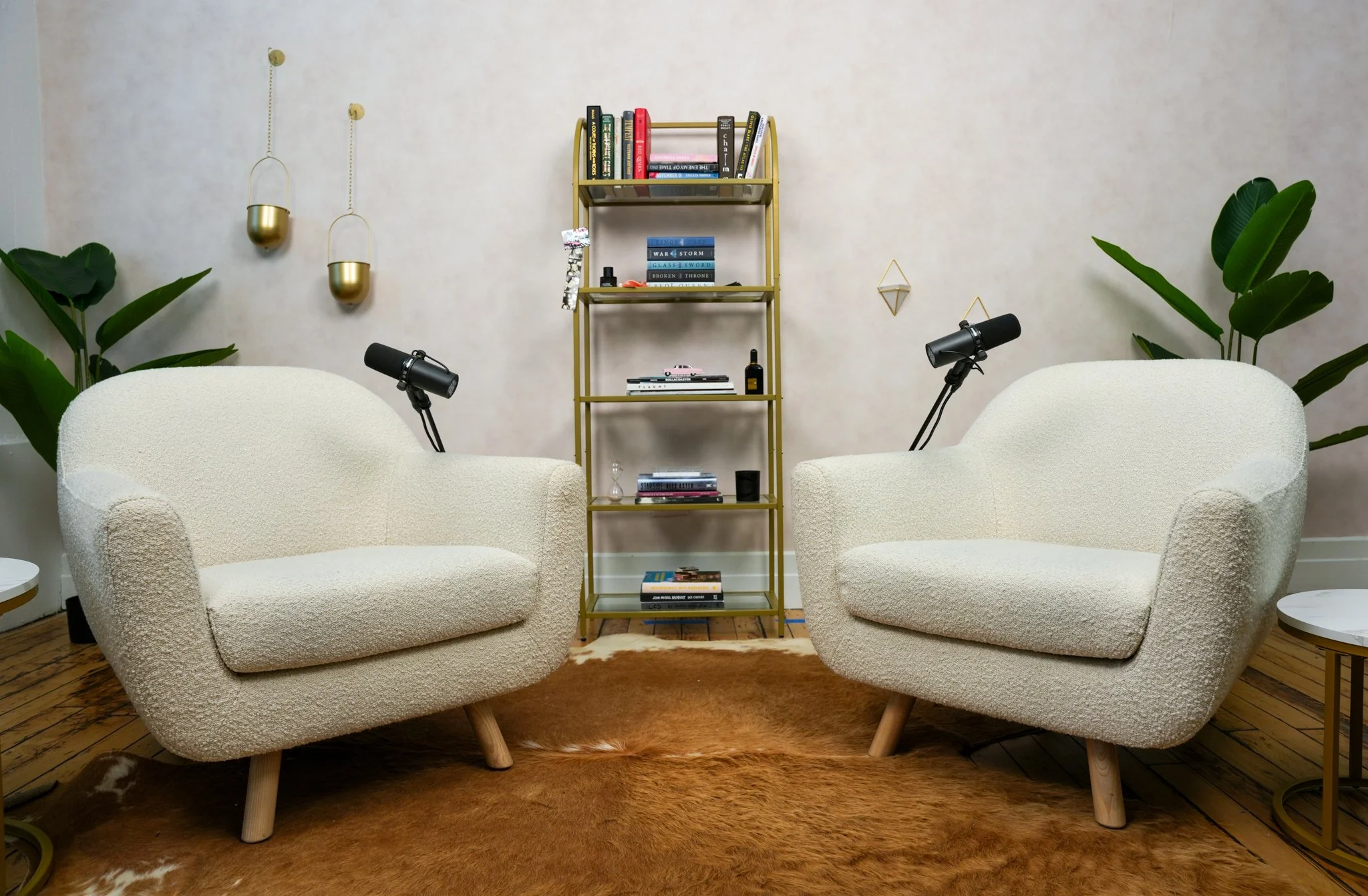 Living room with two white textured armchairs facing each other, a gold bookshelf with books and decor in the background, two black adjustable reading lamps on the chairs, two large green plants, wooden floor, and a brown animal hide rug.