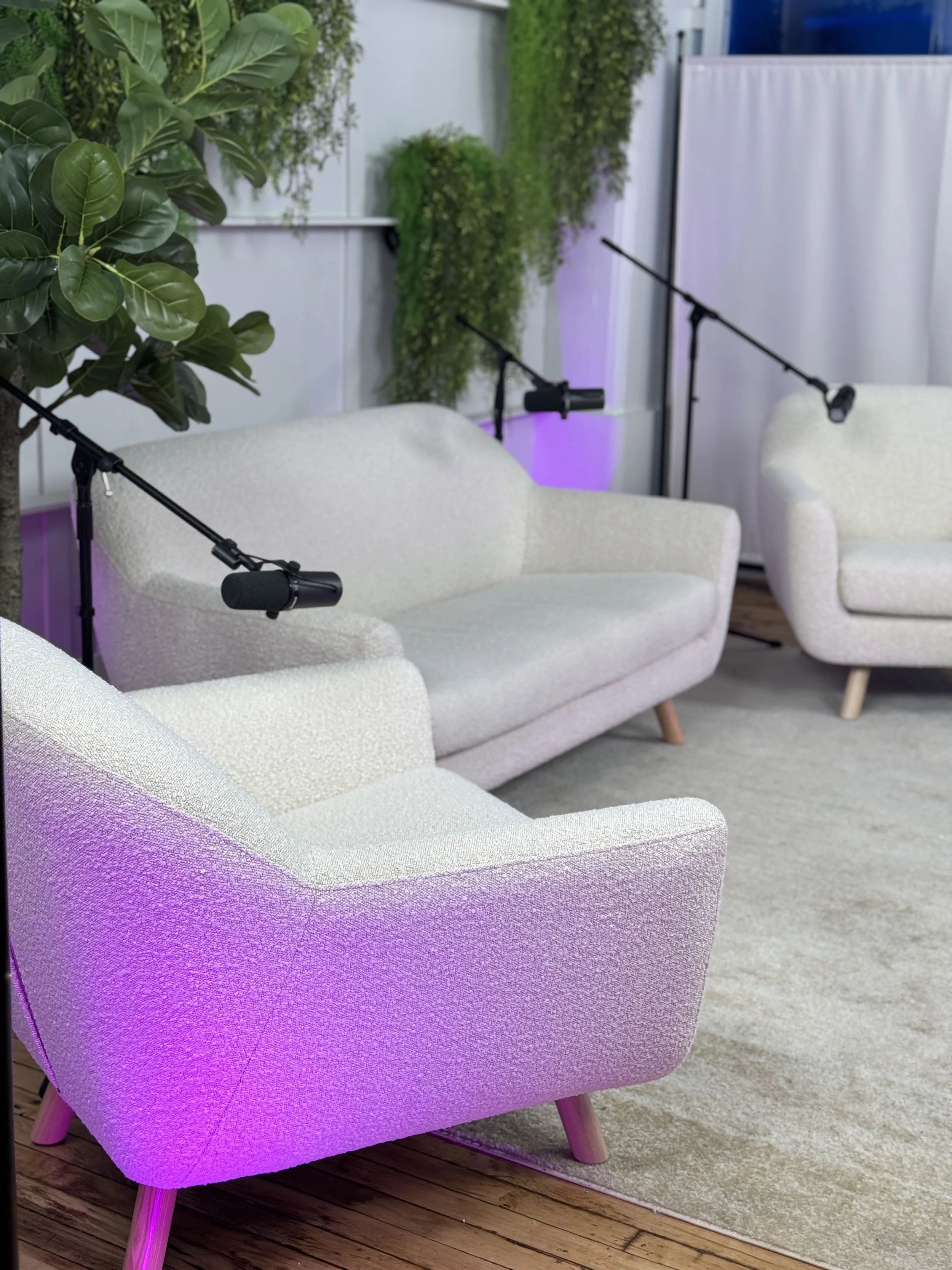 A cozy podcast setup with three white textured sofas, two microphones on adjustable stands, large green plants, and soft purple lighting.