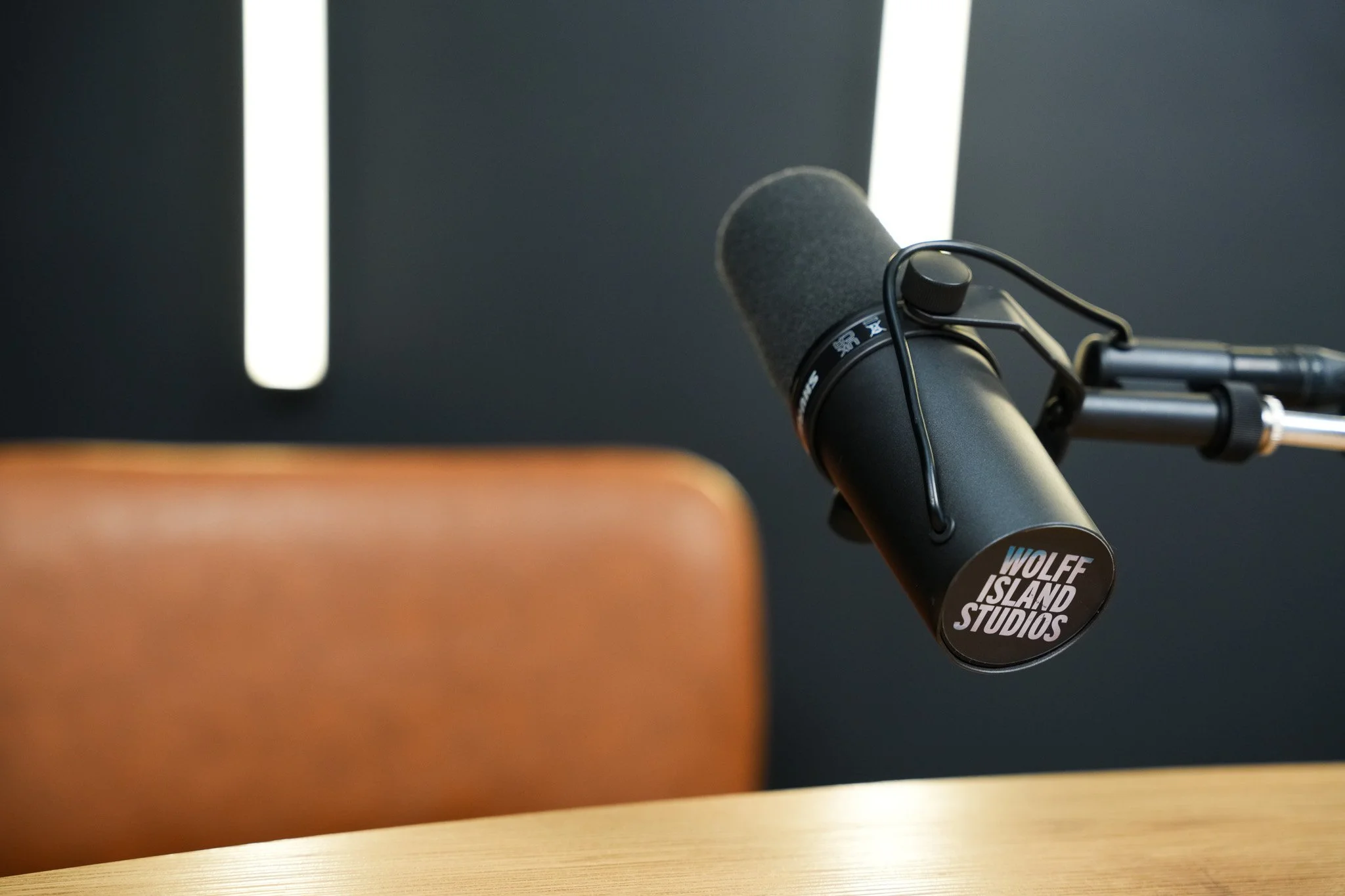 Close-up of a black microphone with a foam windscreen on a stand, with a sign reading 'WOLFE ISLAND STUDIOS', in front of an orange leather chair and a dark wall with white accent lights.
