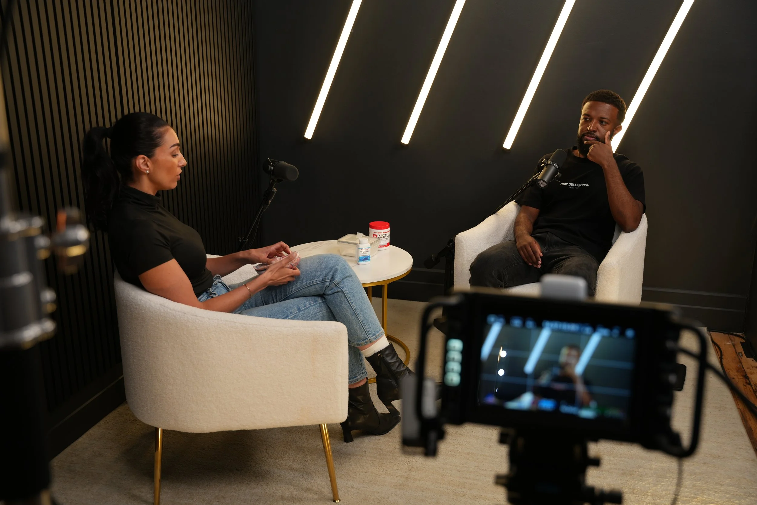 A woman and a man sitting in armchairs during a podcast or interview session, with recording equipment and a camera visible.