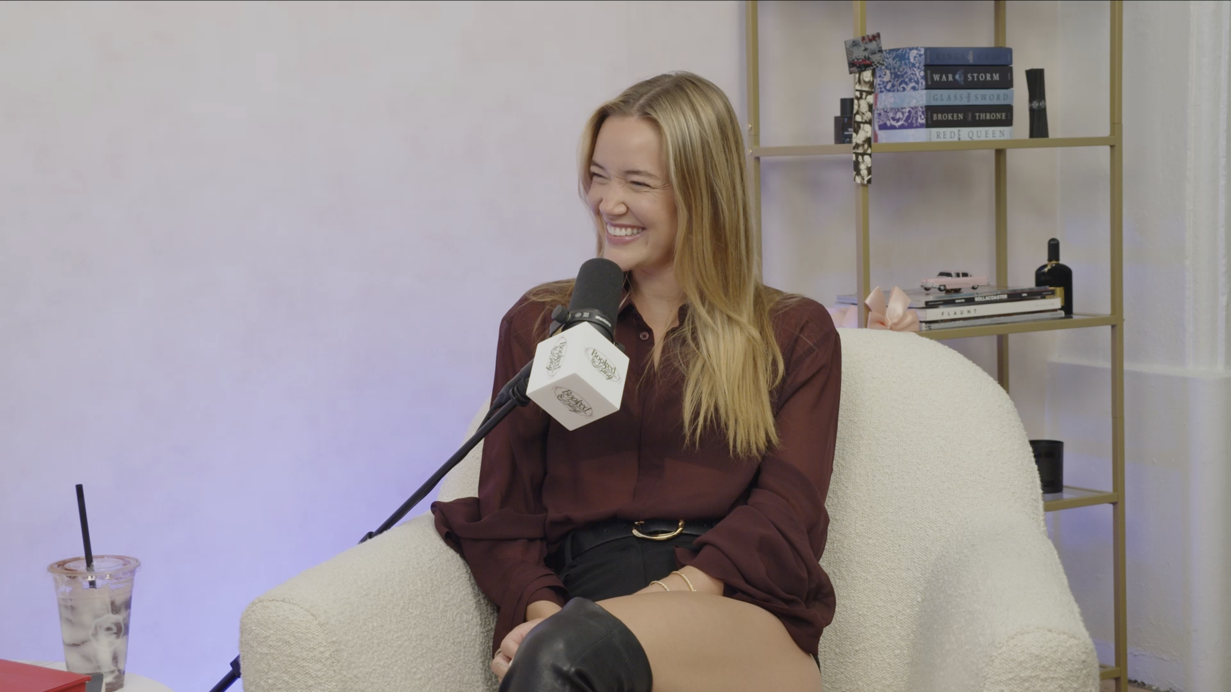 A woman with long blonde hair smiling while speaking into a microphone during an interview in a cozy room with a bookshelf and decorative items in the background.