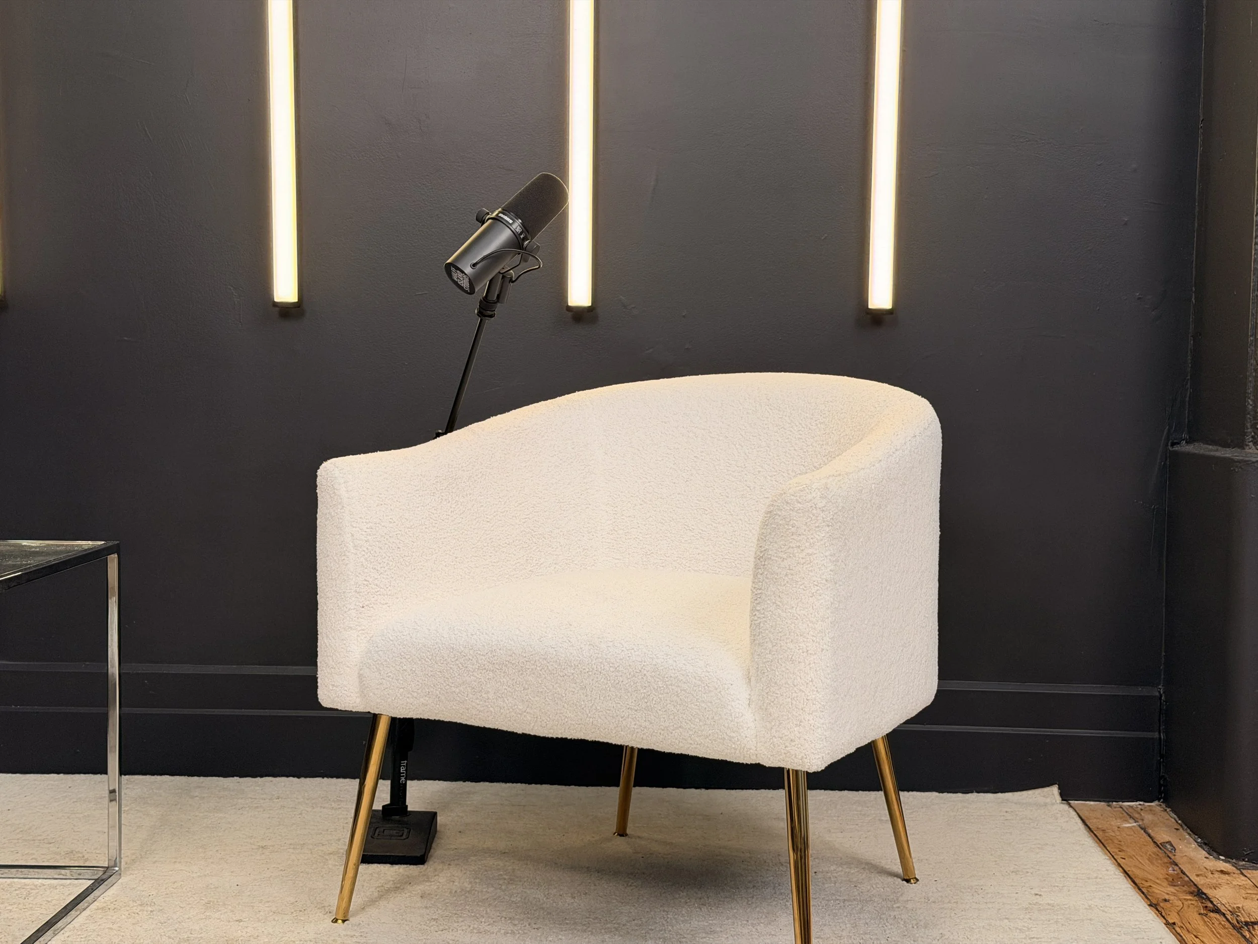A white textured armchair with gold legs, accompanied by a black microphone on a stand, set against a black wall with vertical light fixtures.