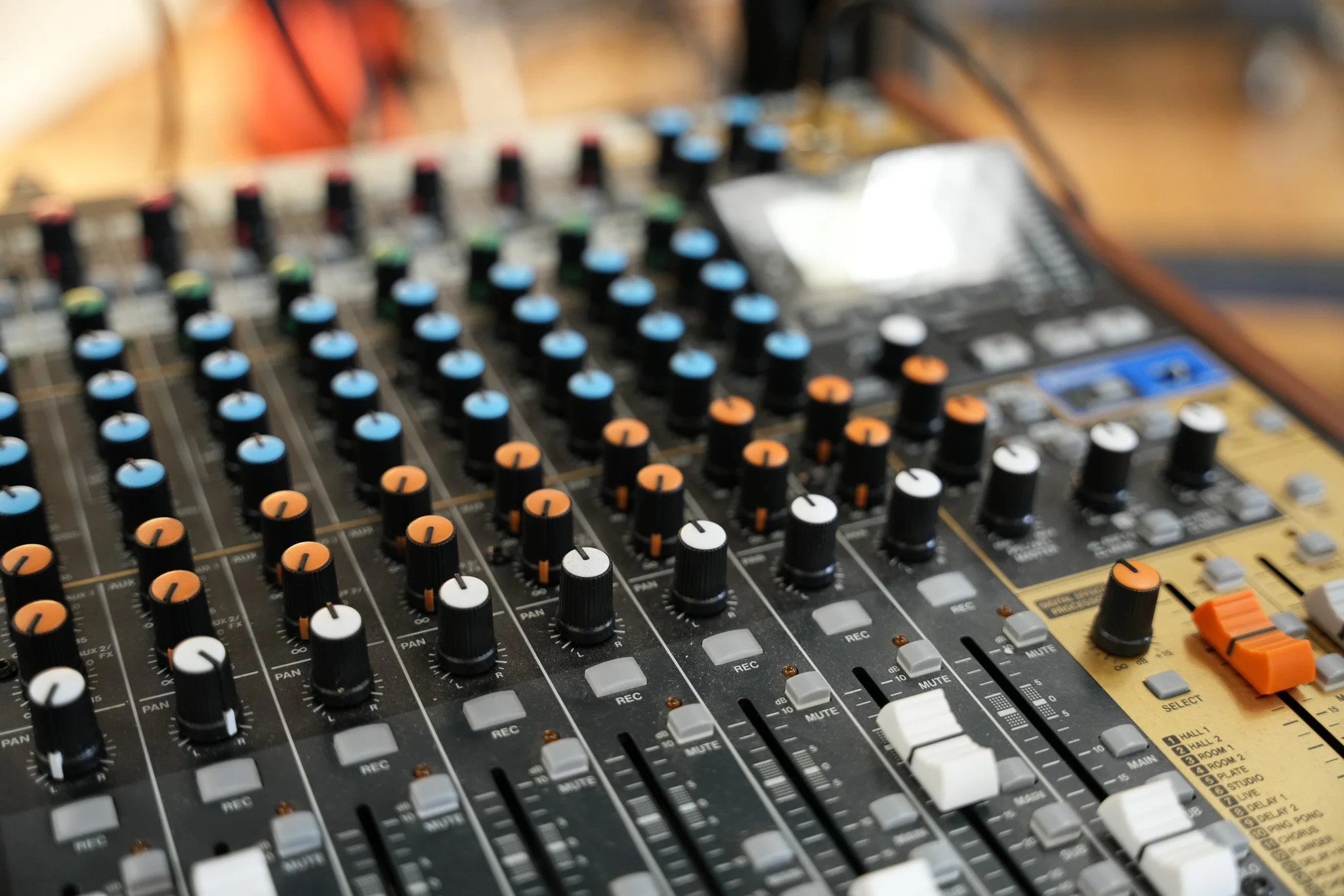 A close-up view of a professional audio mixing console with numerous knobs, sliders, and buttons in black, white, blue, and orange colors.