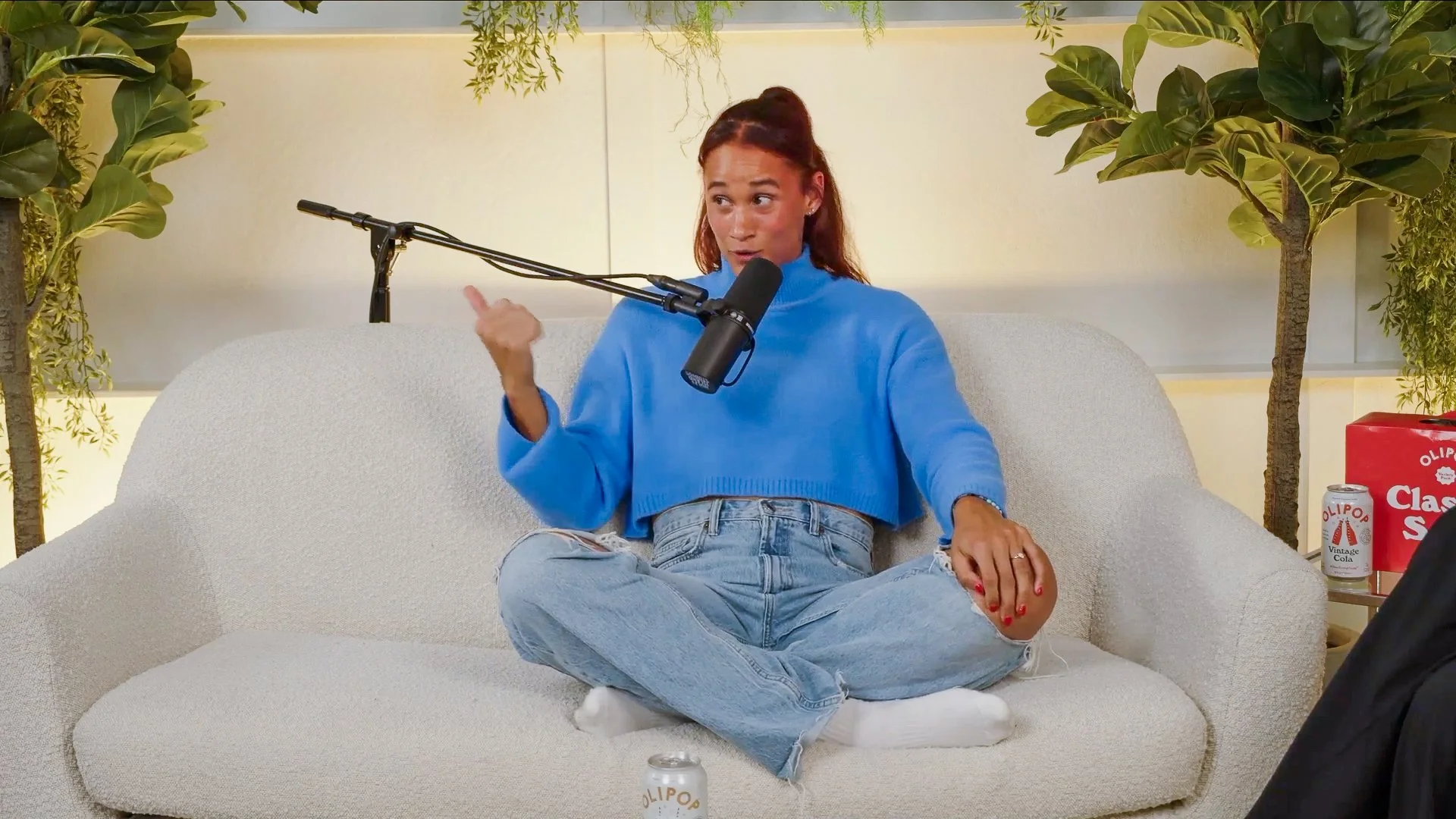 A woman with red hair wearing a blue sweater and ripped jeans sits on a white couch with her legs crossed. She is speaking into a microphone attached to a stand, gesturing with her right hand. There are green plants behind her and some snacks and dri