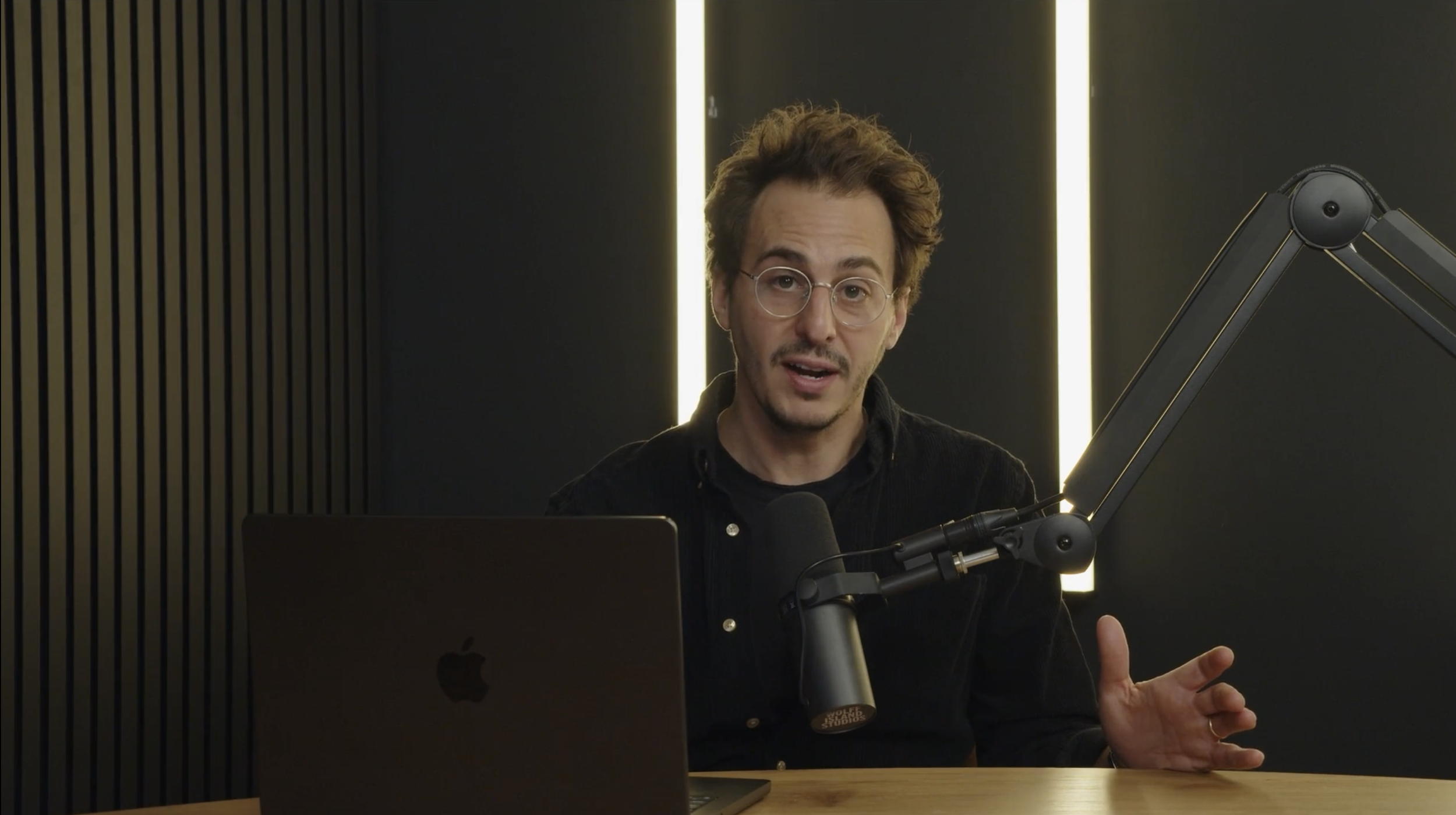 A man with glasses and messy hair speaks into a microphone in a recording studio or podcast setup, with a laptop in front of him and dark background with vertical light strips.