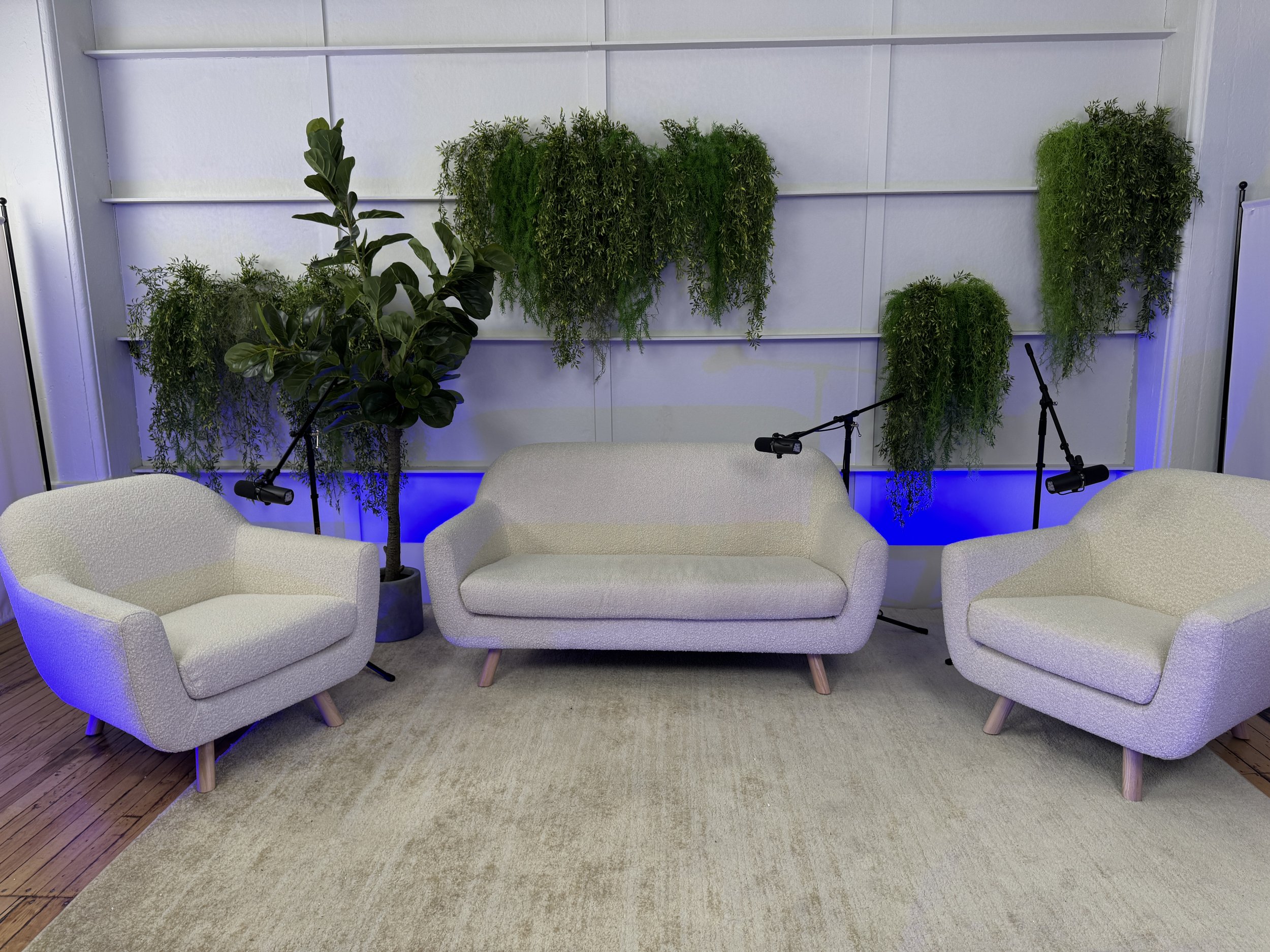 A modern, cozy indoor living room setup with four light-colored, cushioned furniture pieces — one sofa and three armchairs — arranged on a beige rug. Behind the furniture is a white wall decorated with a vertical garden of lush green plants and blue 