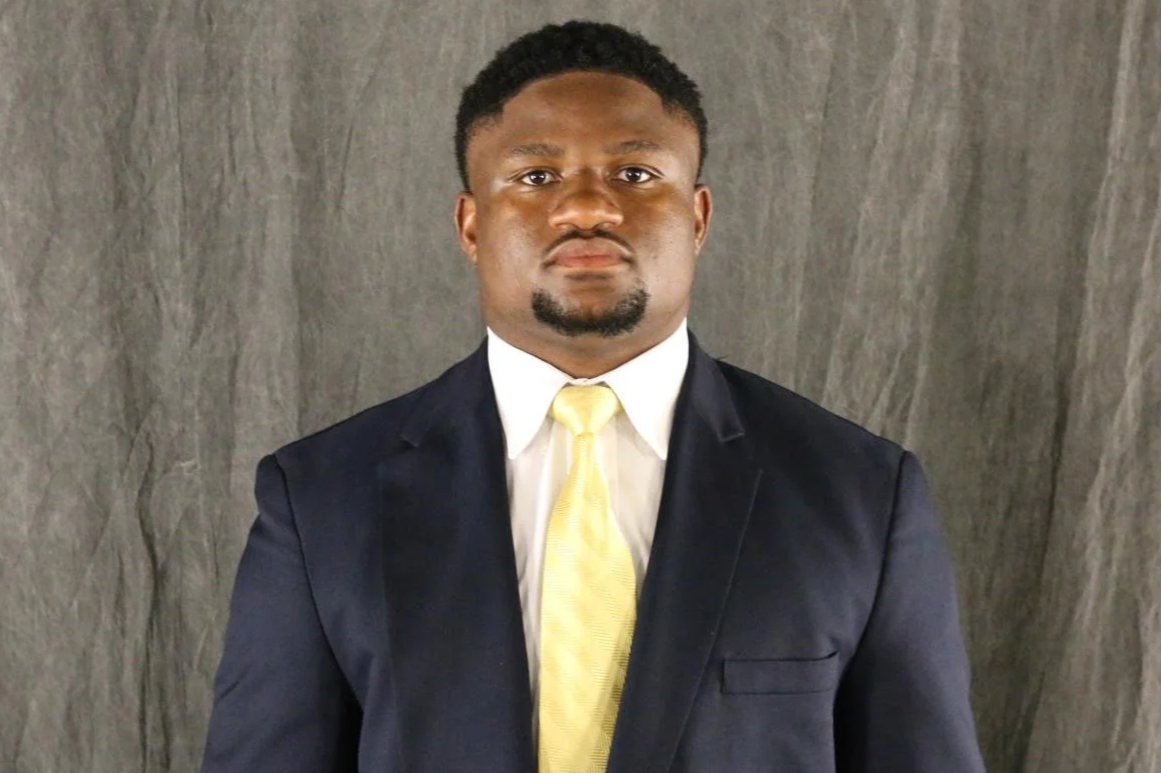 A man with glasses, dressed in a dark blue suit and white shirt, smiling against a plain dark background.