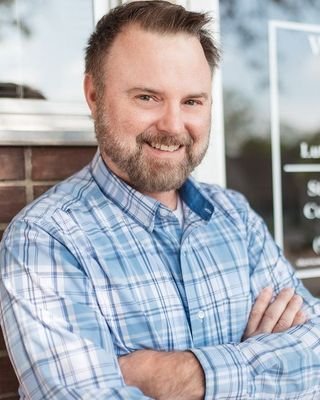 A man with a beard and short hair smiling outdoors, wearing a blue plaid shirt, with a building and glass window in the background.