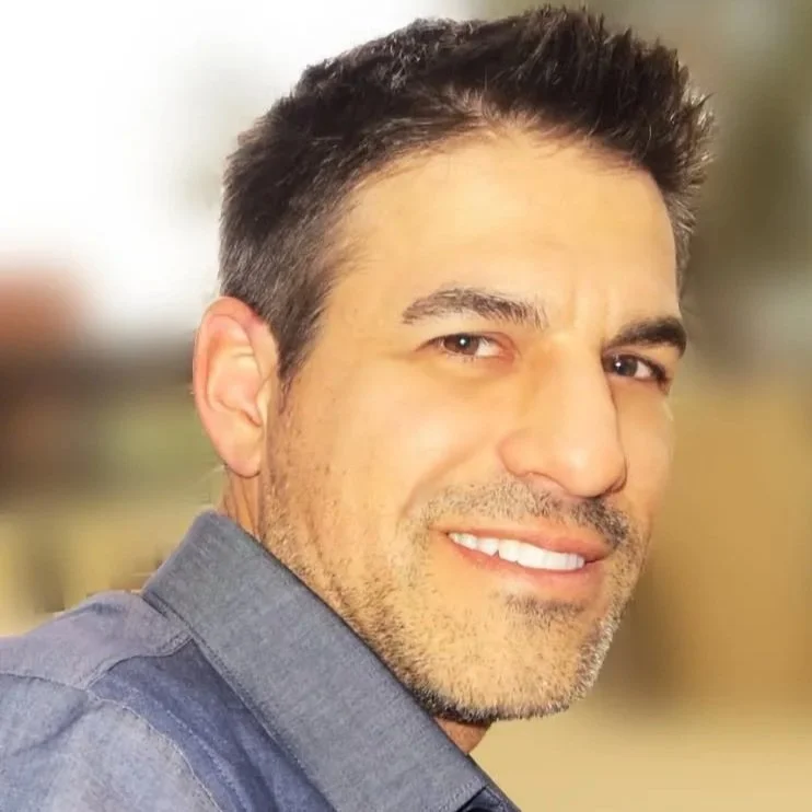 Close-up of a smiling man with short dark hair and stubble, wearing a gray shirt, in an indoor setting with blurred background.
