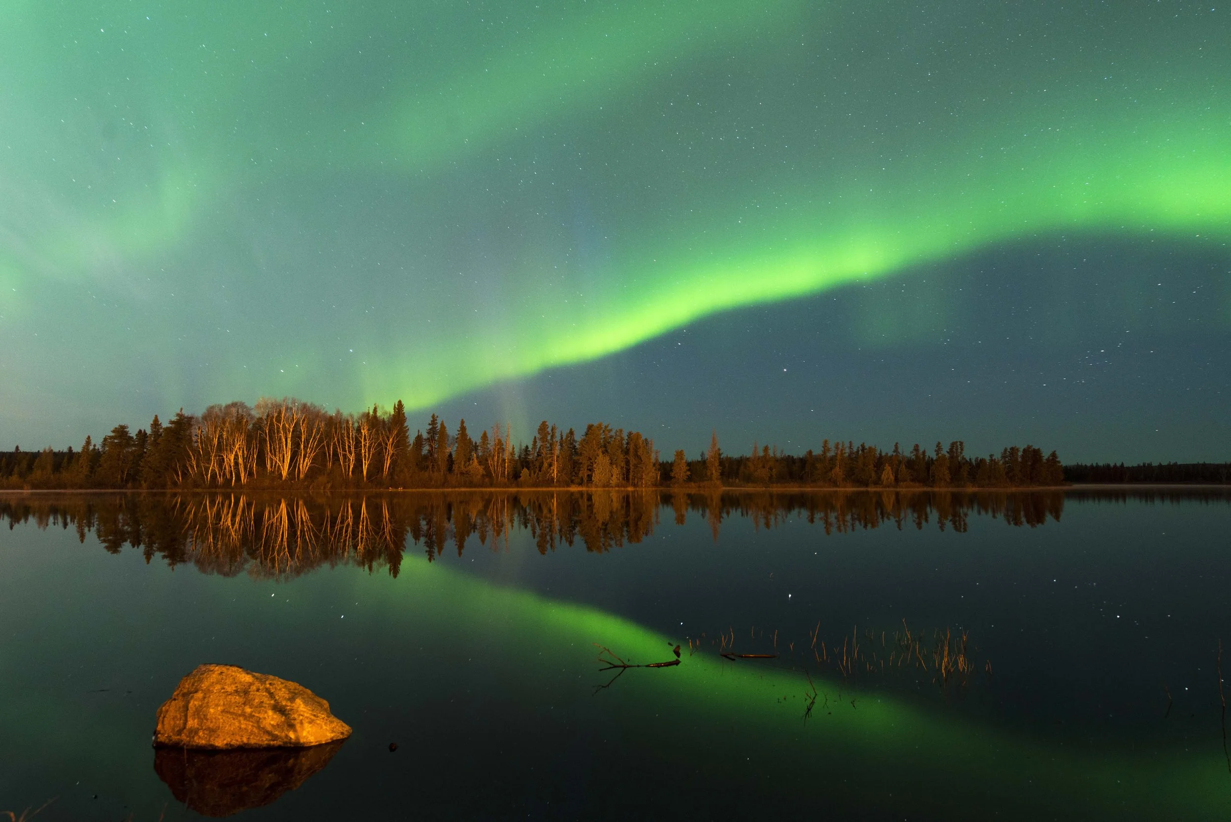 Northern lights or aurora borealis in green colors over a lake with a forest and a large rock reflected in the water.