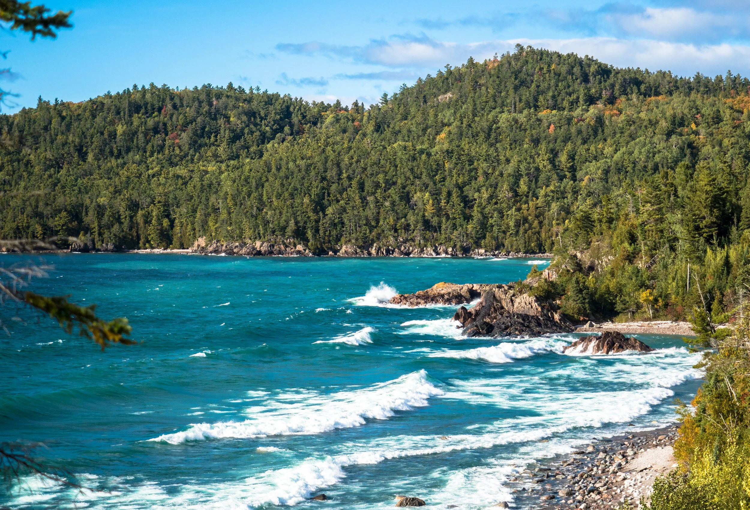 A scenic coastal landscape with a forested hillside leading to a rocky shoreline and turquoise ocean water under a partly cloudy sky.