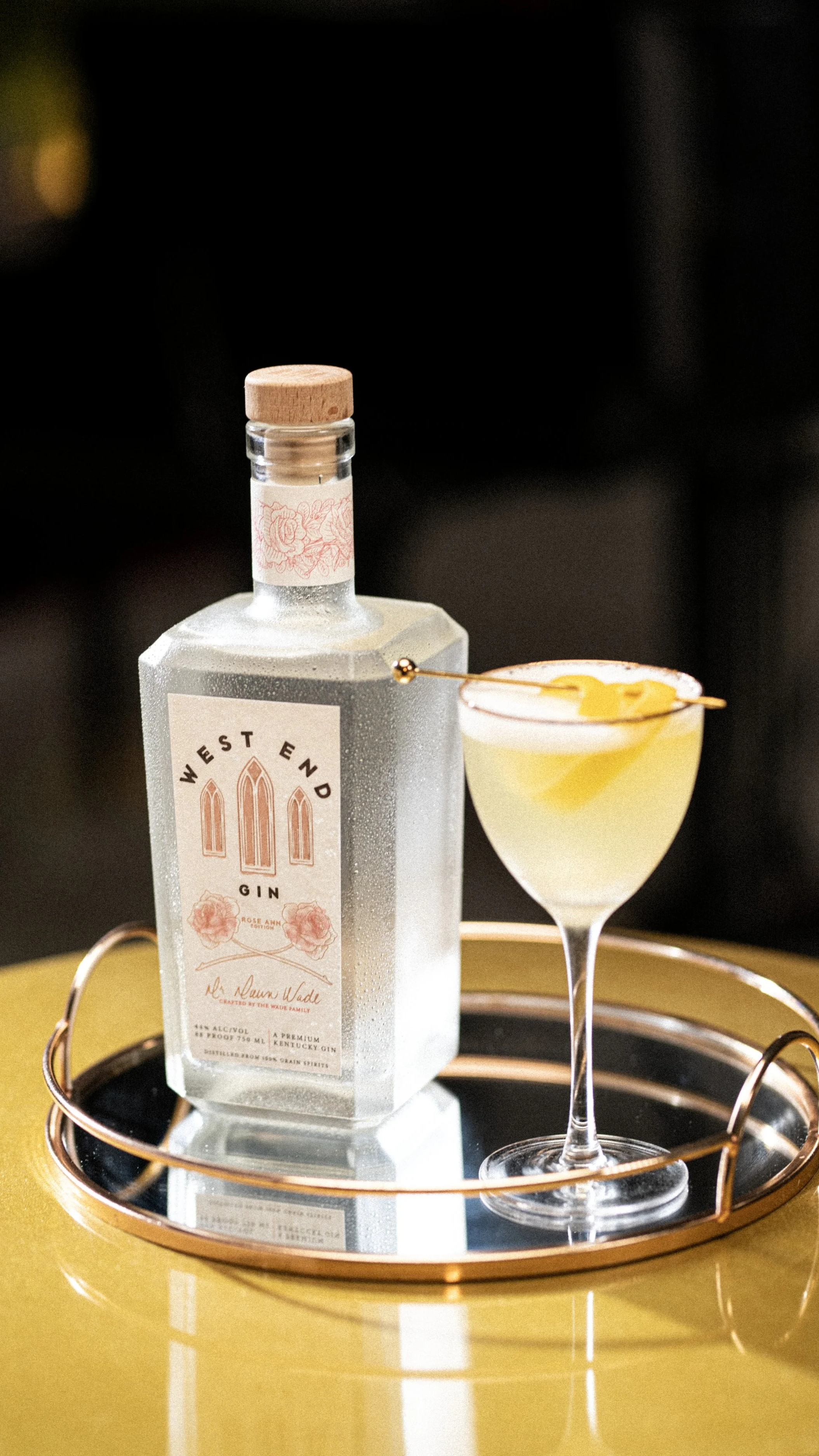 Bottle of gin and cocktail glass on a brass tray with lemon slices in a wooden bowl.