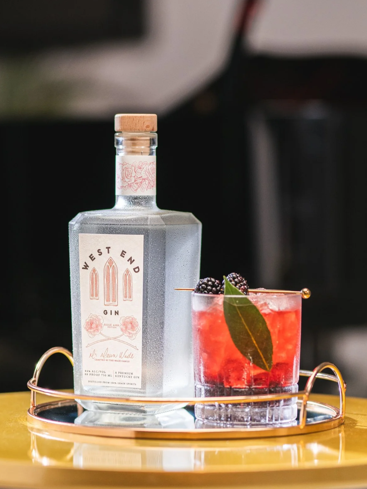 Red cocktail with blackberry and mint garnish, on a serving tray with a square gin bottle