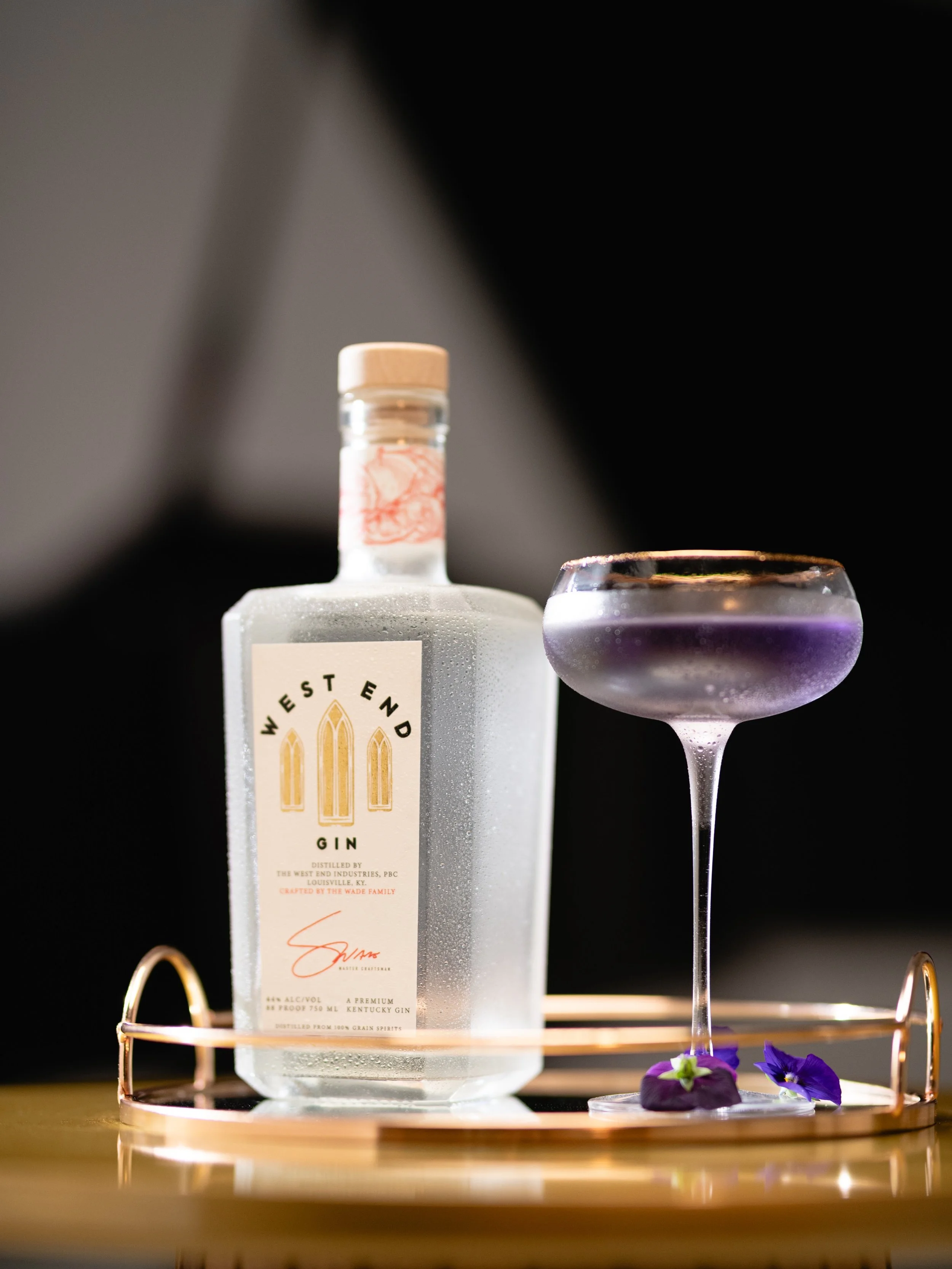 Purple cocktail with floral garnish on a serving tray with a square gin bottle