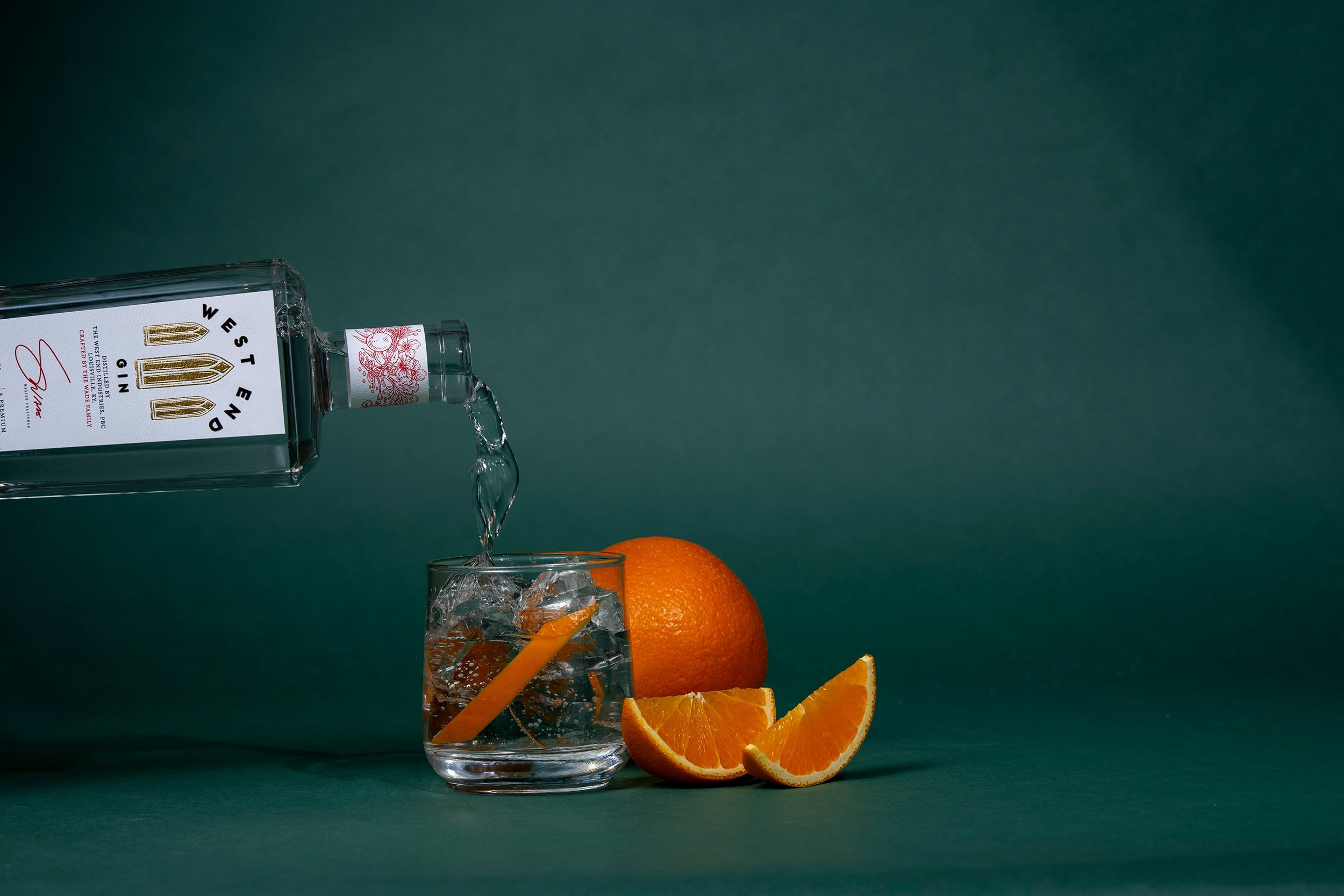 A bottle of Western gin tilted pouring clear liquid into a glass with ice and orange slices, with a whole orange and orange wedges beside the glass on a green background.