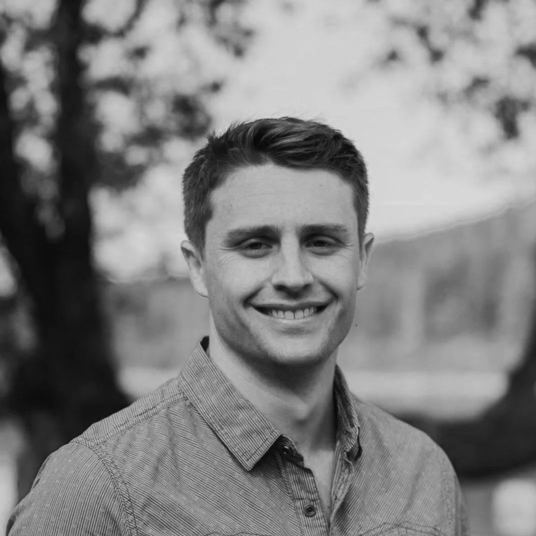 Black and white portrait of a young man smiling outdoors, wearing a casual button-up shirt, with blurry trees in the background.