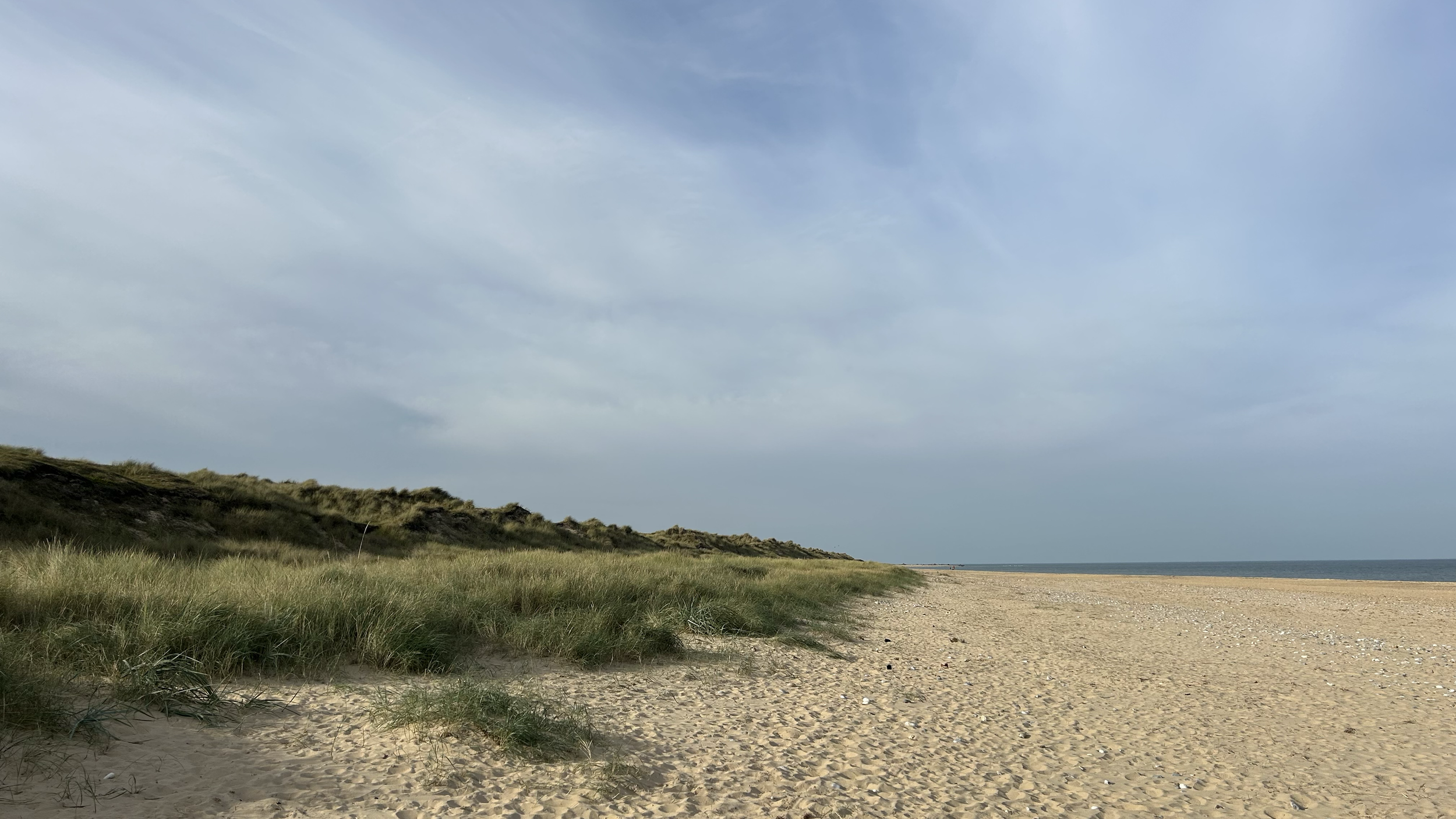 Empty sandy beach with green grassy dunes on the left and calm ocean in the distance, under a partly cloudy sky.