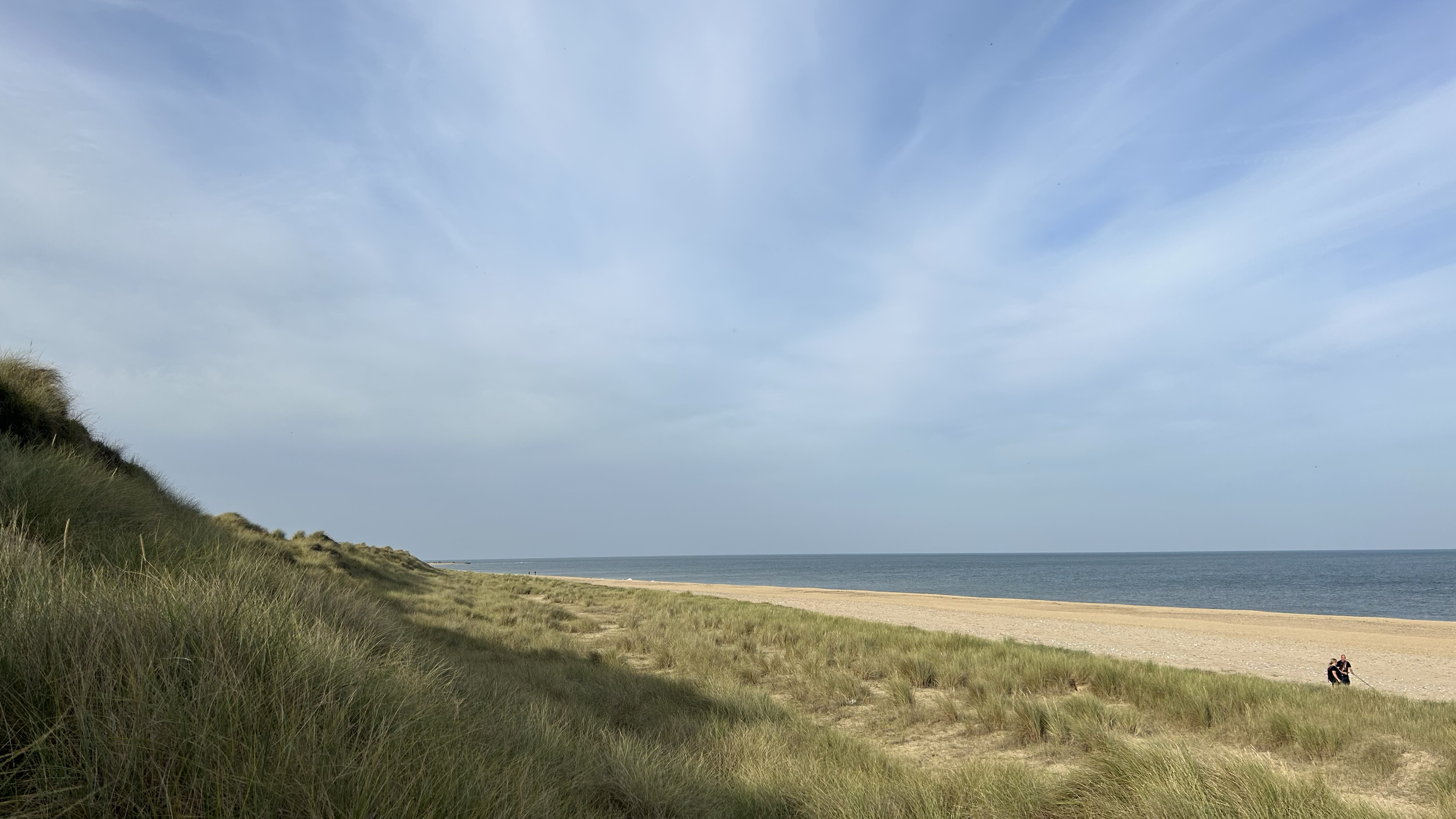 A sandy beach with grassy dunes on the left and a calm ocean under a partly cloudy sky. Two people are sitting in the distance on the right.