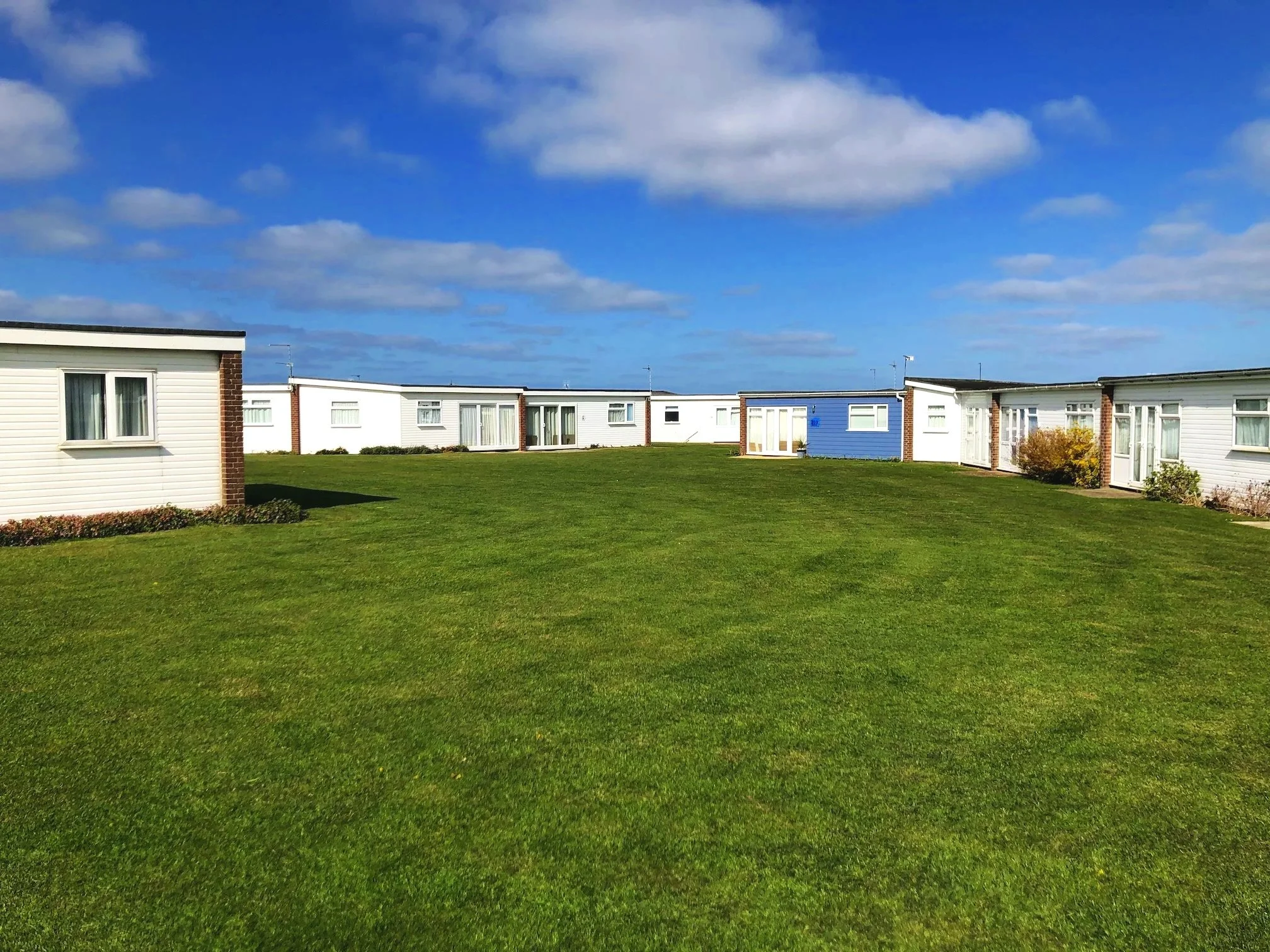 Row of chalets with different coloured siding, surrounded by green grass and a blue sky with scattered clouds.
